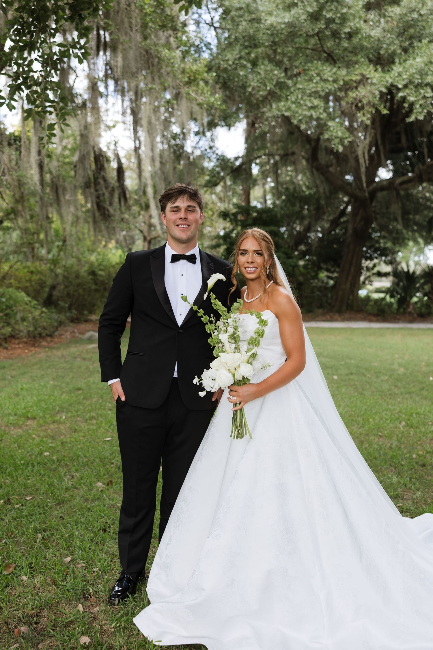 Bride and groom posing outdoors under Spanish moss trees, bride in white ballgown holding white bouquet, groom in black tuxedo.