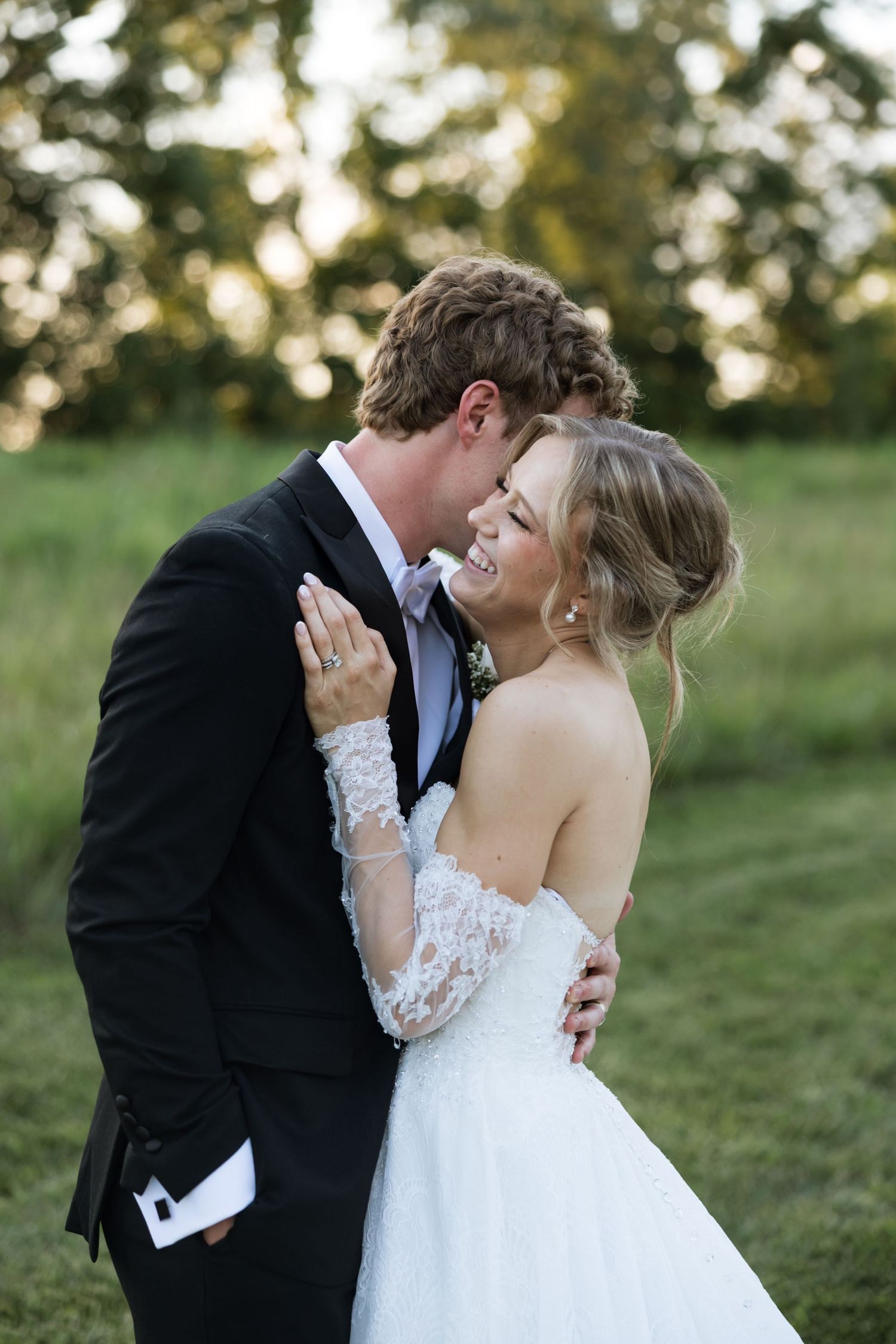 Groom in black tuxedo embraces smiling bride in lace off-shoulder gown outdoors at sunset.