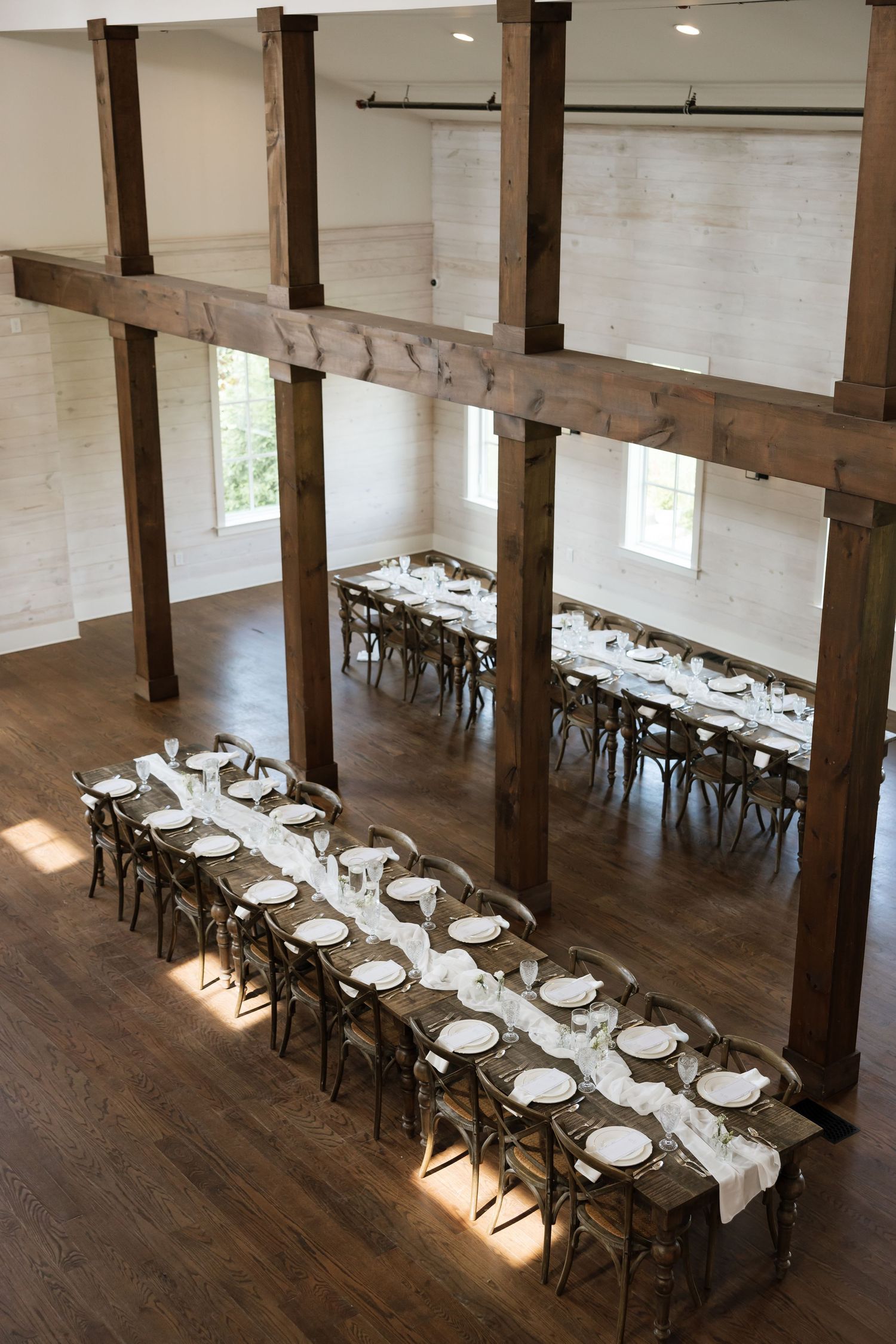 Elegant barn wedding reception with long farmhouse tables, white place settings, and rustic wooden beams on hardwood floors.