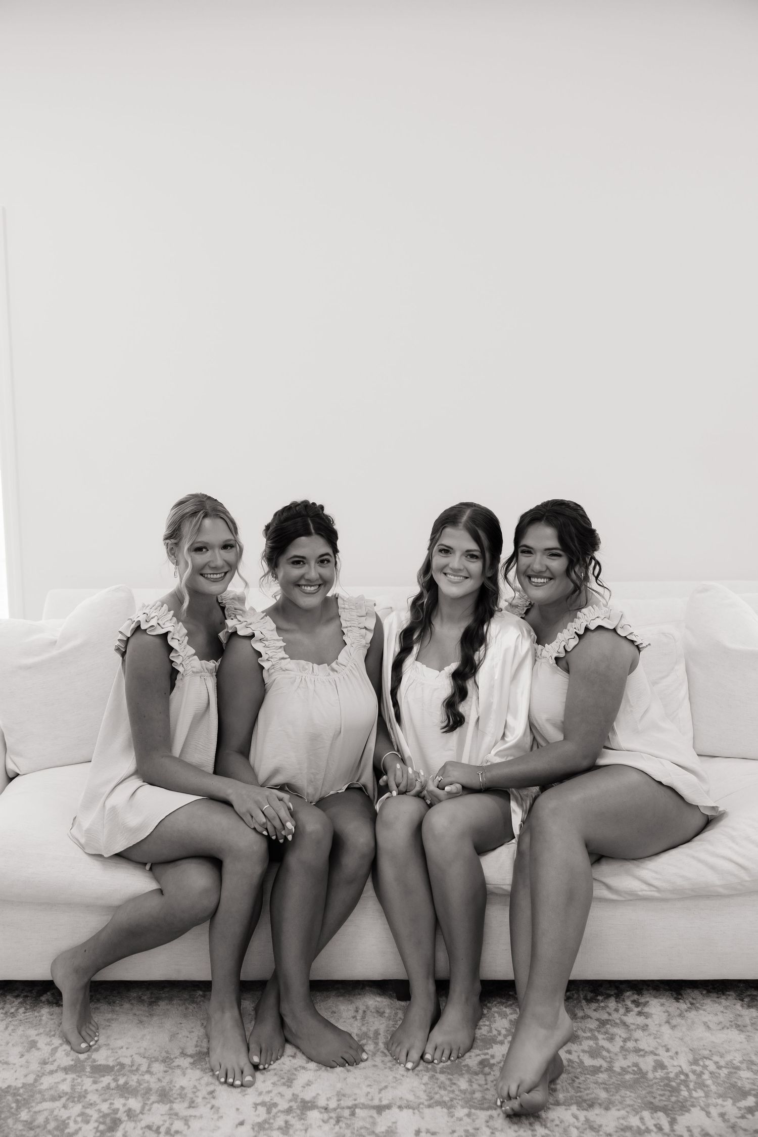 Four bridesmaids in matching robes sit barefoot on a white couch, smiling together in a black and white photo.