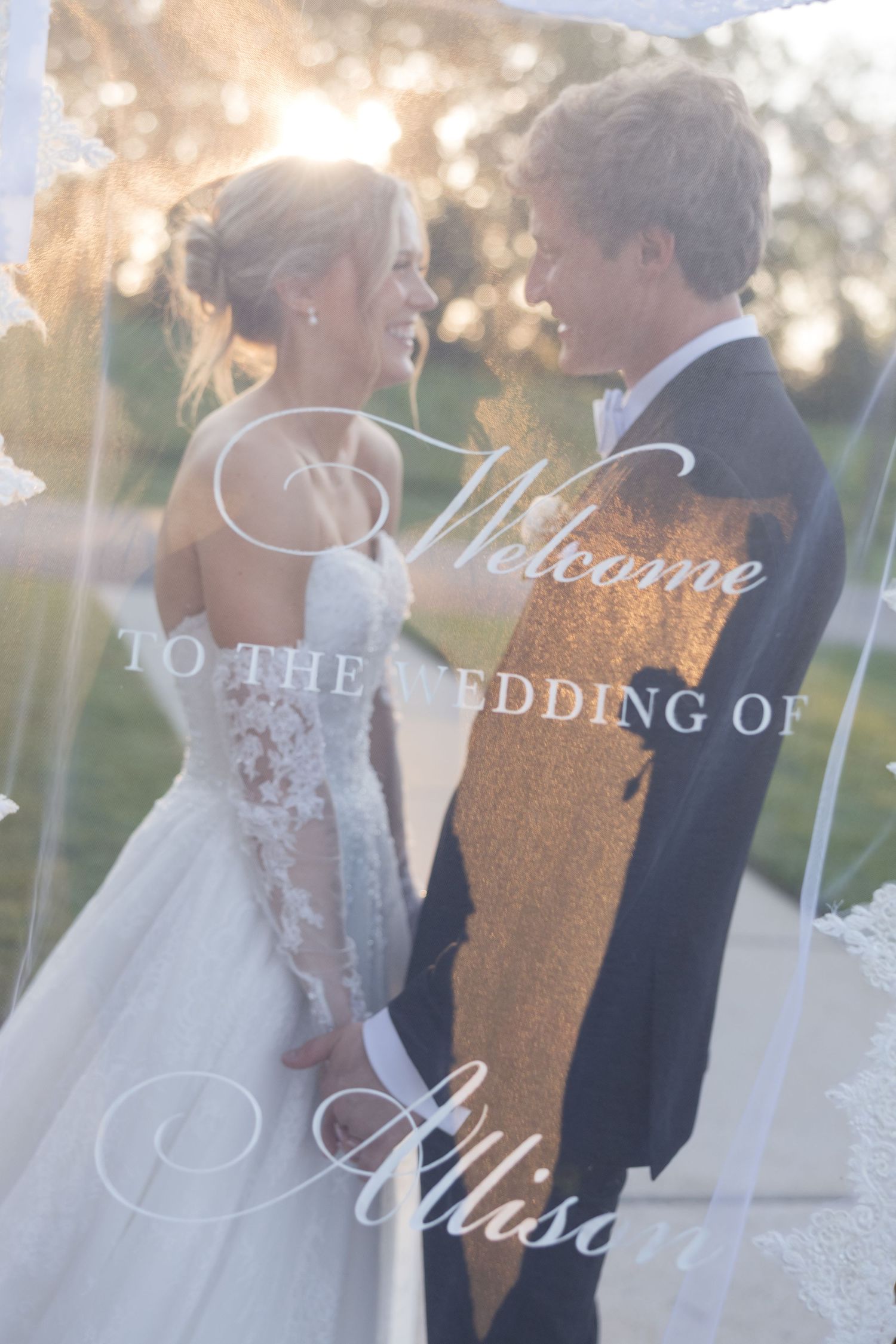 Bride and groom smiling at each other behind a wedding welcome sign reading Welcome to the Wedding of Allison.