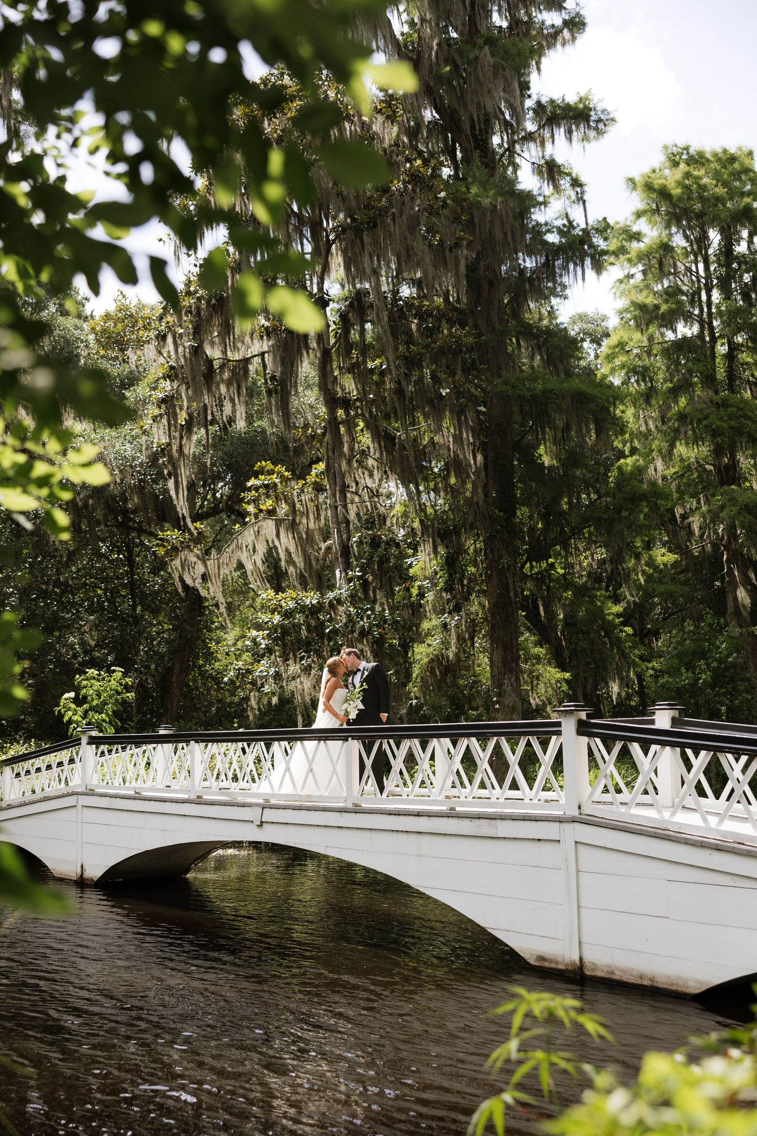 Couple sharing a romantic kiss on a white ornamental bridge surrounded by lush greenery and Spanish moss-draped trees.