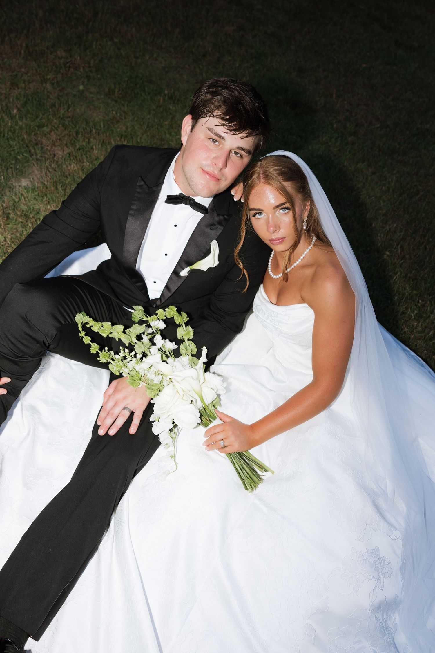 Bride and groom sitting on grass at night, she in white gown with bouquet, he in black tuxedo.