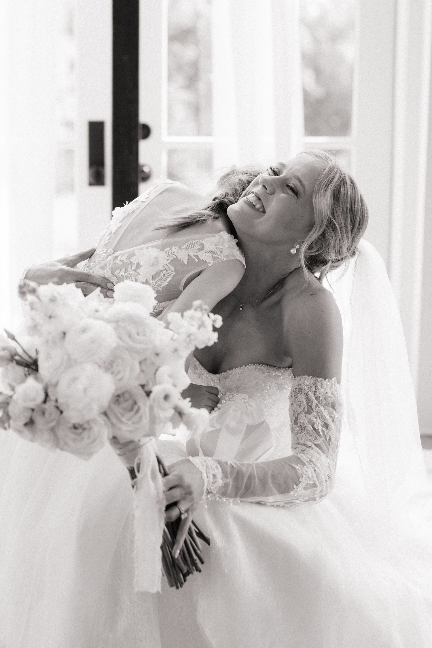 Black and white photo of a joyful bride in lace gown holding a white floral bouquet, laughing with eyes closed.