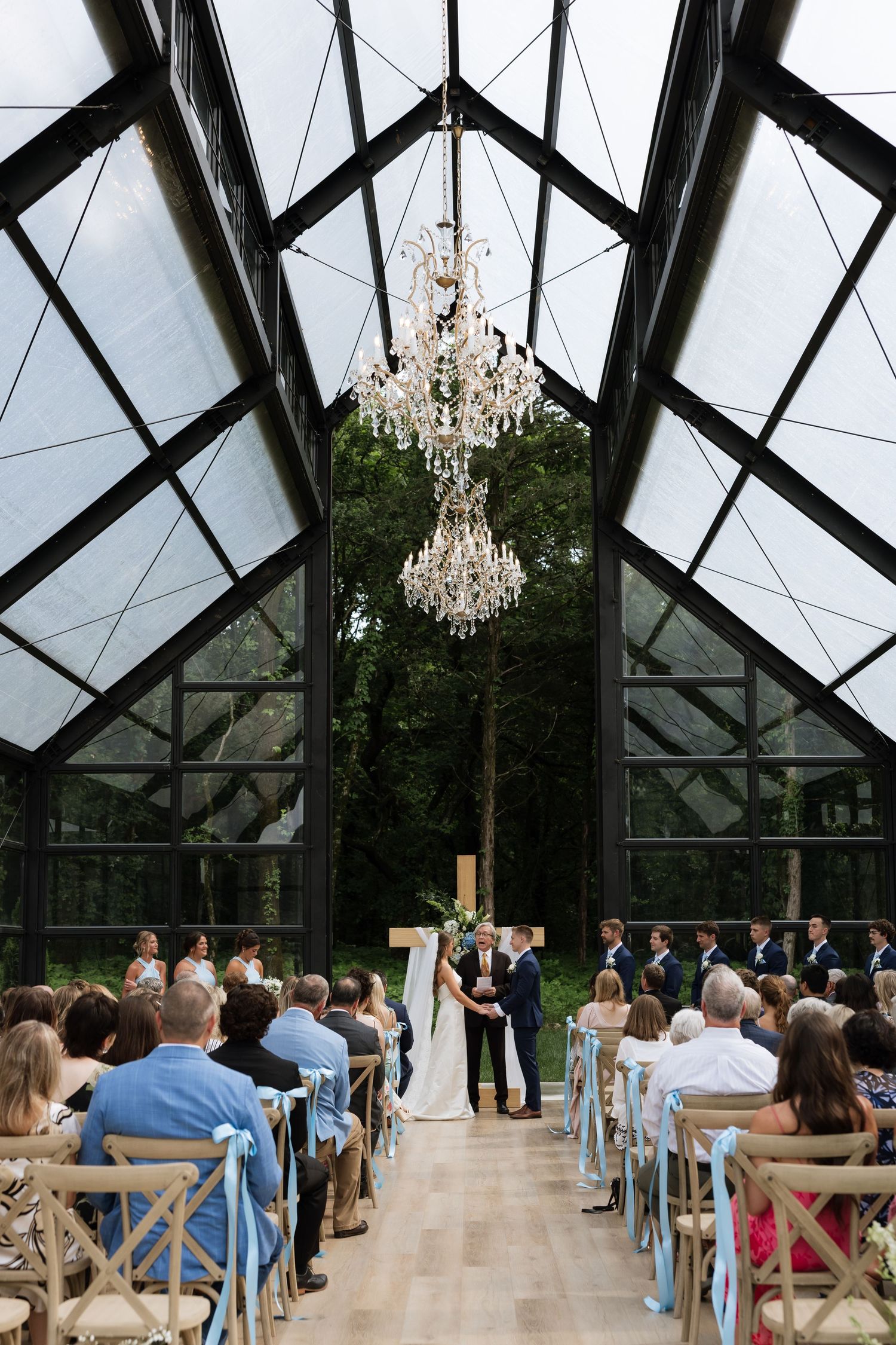Wedding ceremony in a glass greenhouse venue with chandeliers, guests seated in wooden chairs along a floral aisle.