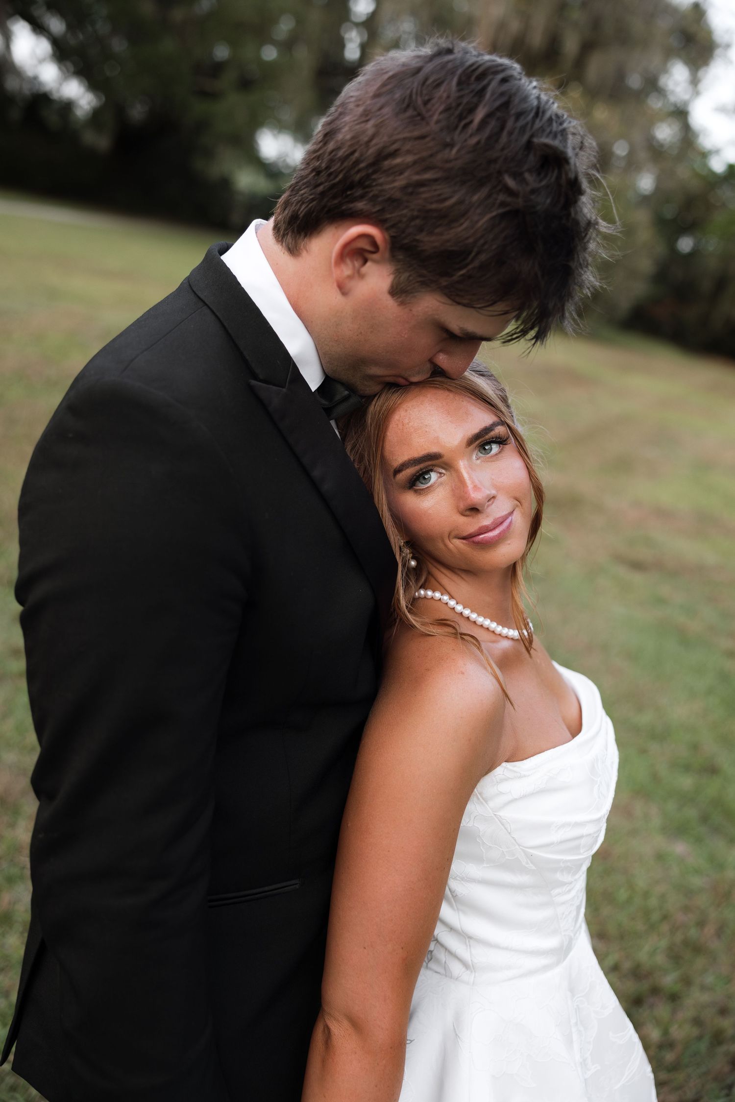 Bride in white strapless gown with pearl necklace leans against groom in black tuxedo outdoors.