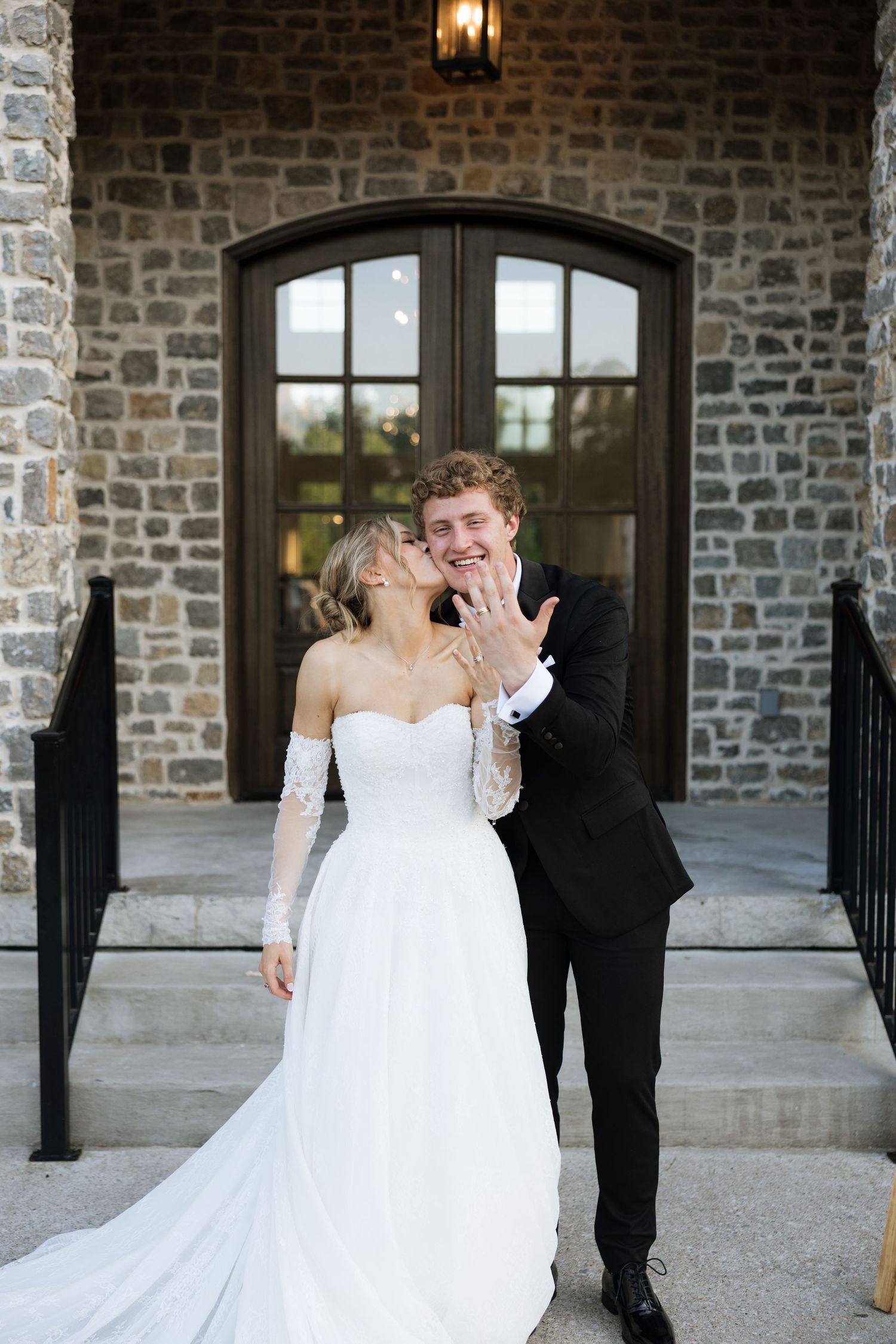 Smiling groom shows off wedding ring while bride kisses his cheek outside stone venue entrance.