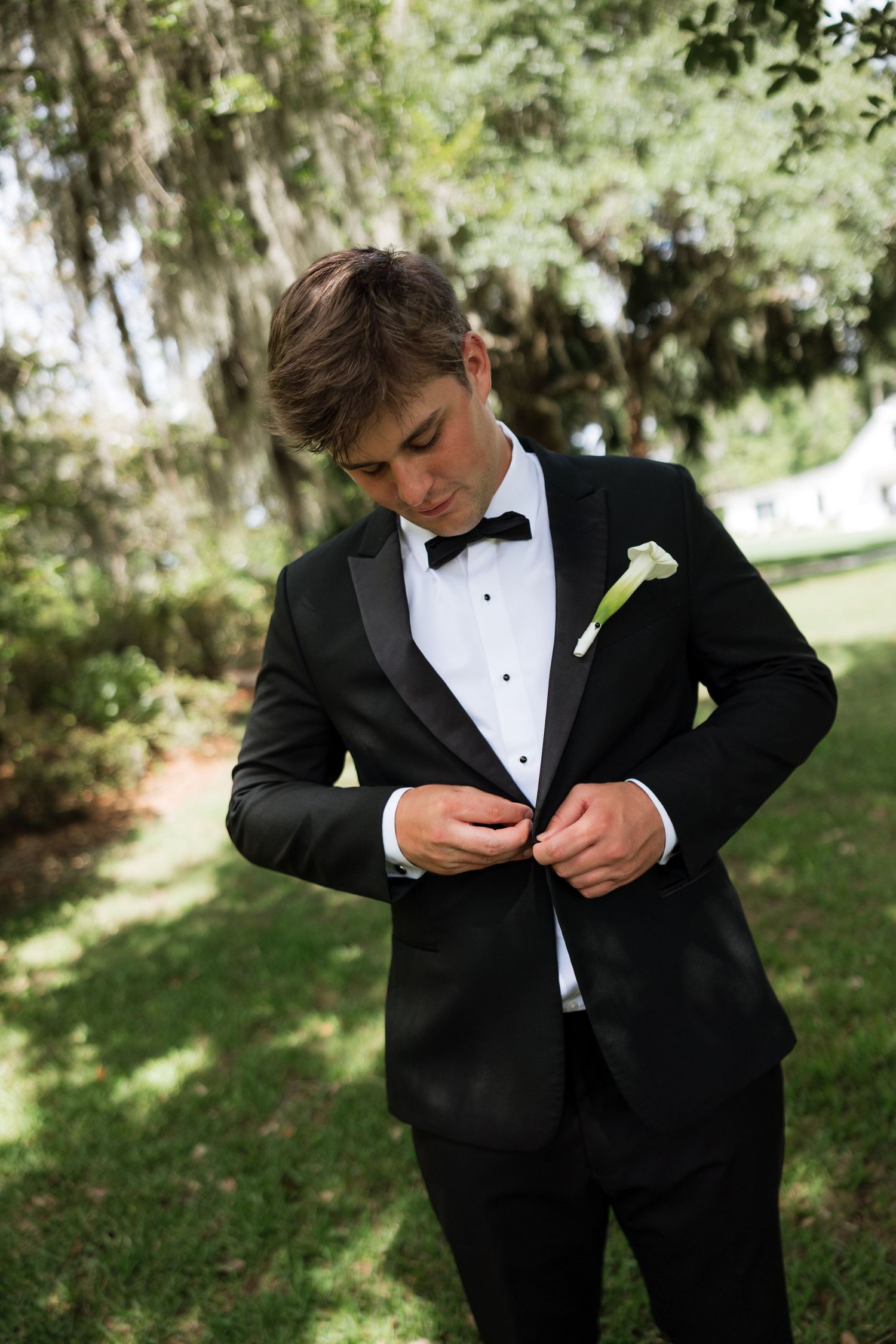Young groom in black tuxedo with white calla lily boutonniere buttoning jacket outdoors under moss-draped trees.