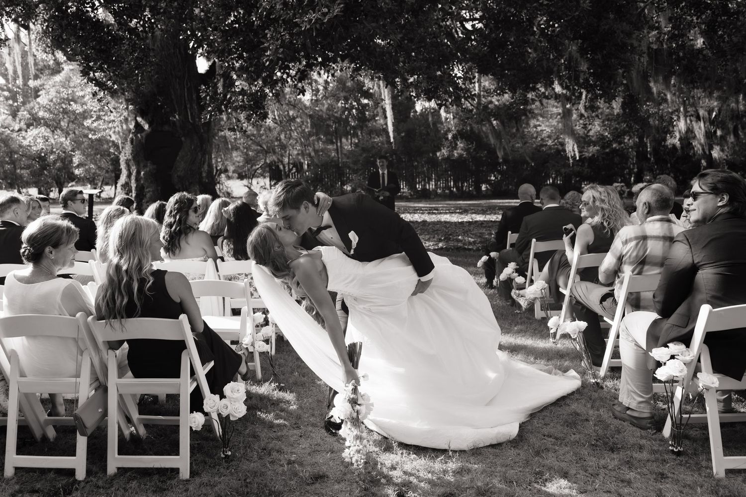 Black and white photo of a couple sharing a kiss at an outdoor wedding ceremony surrounded by seated guests.