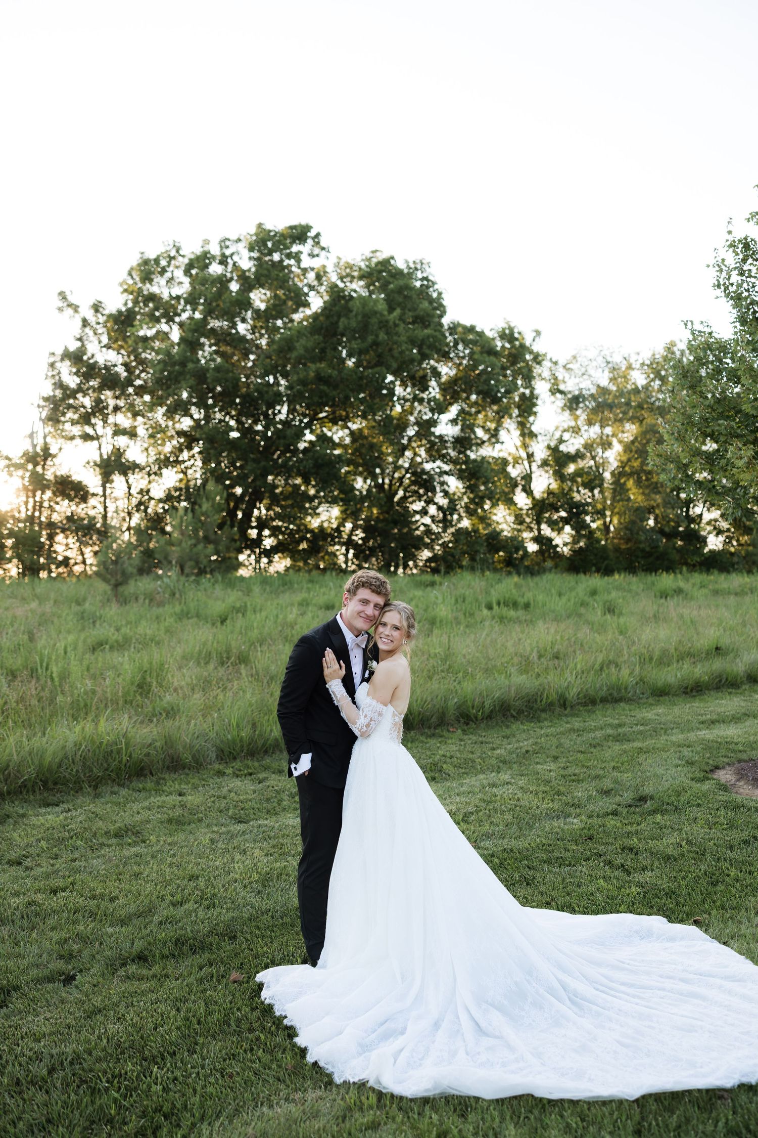 Bride and groom share romantic sunset portrait on lush green lawn, bride wearing long cathedral train wedding gown.