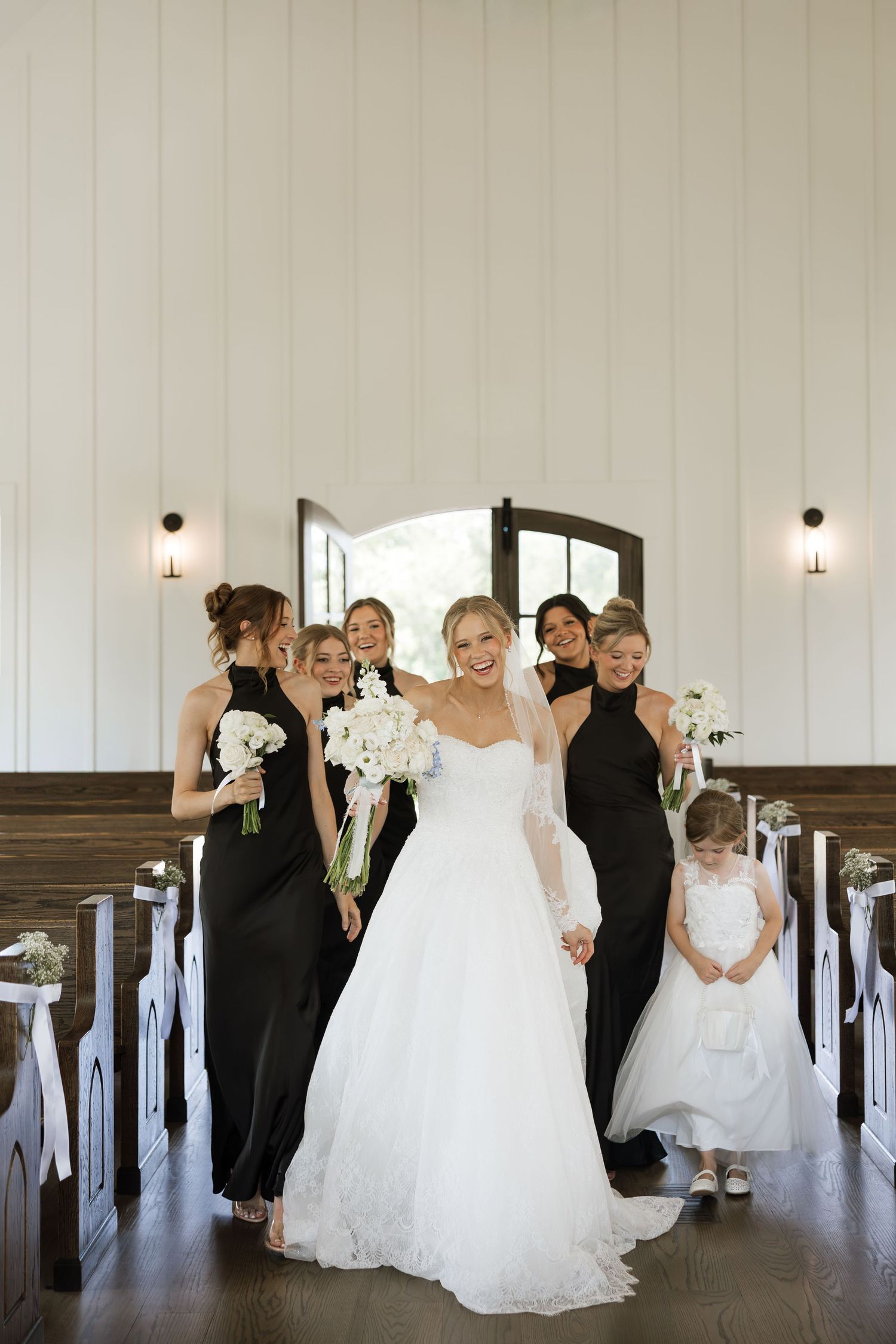 Bride in white gown with bridesmaids in black dresses and flower girl walking down chapel aisle.