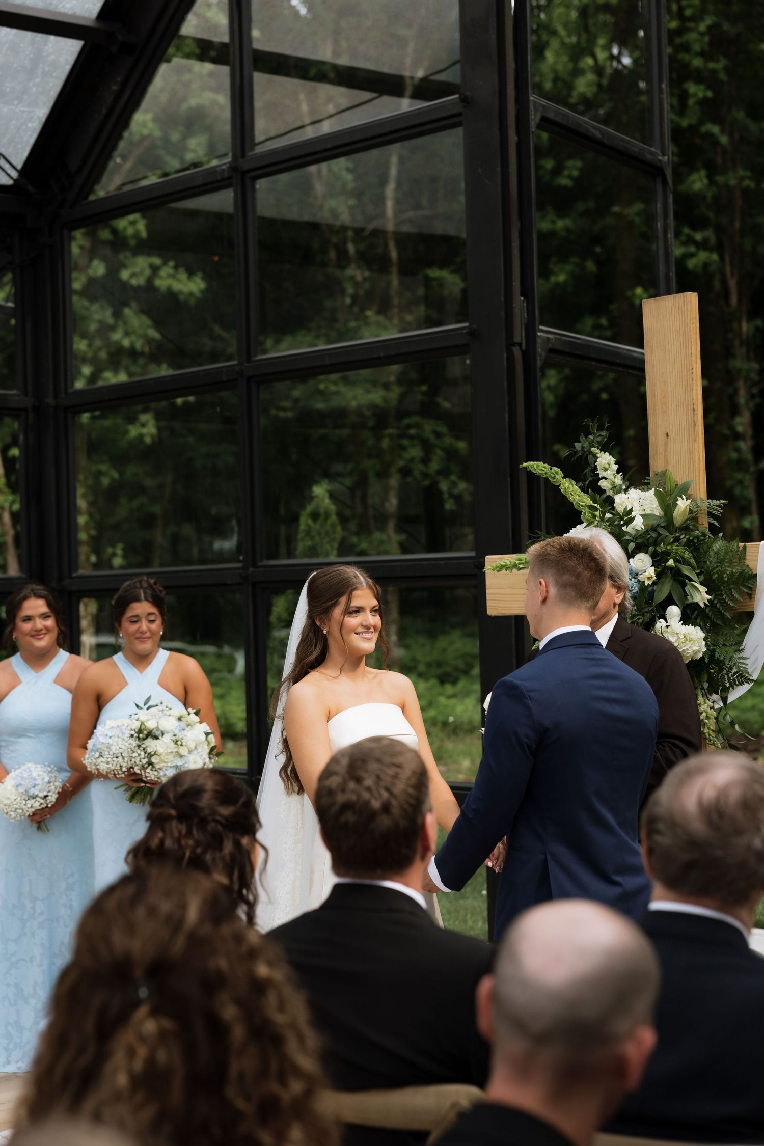 Bride smiling at groom during outdoor wedding ceremony with bridesmaids in blue dresses and floral arrangements.