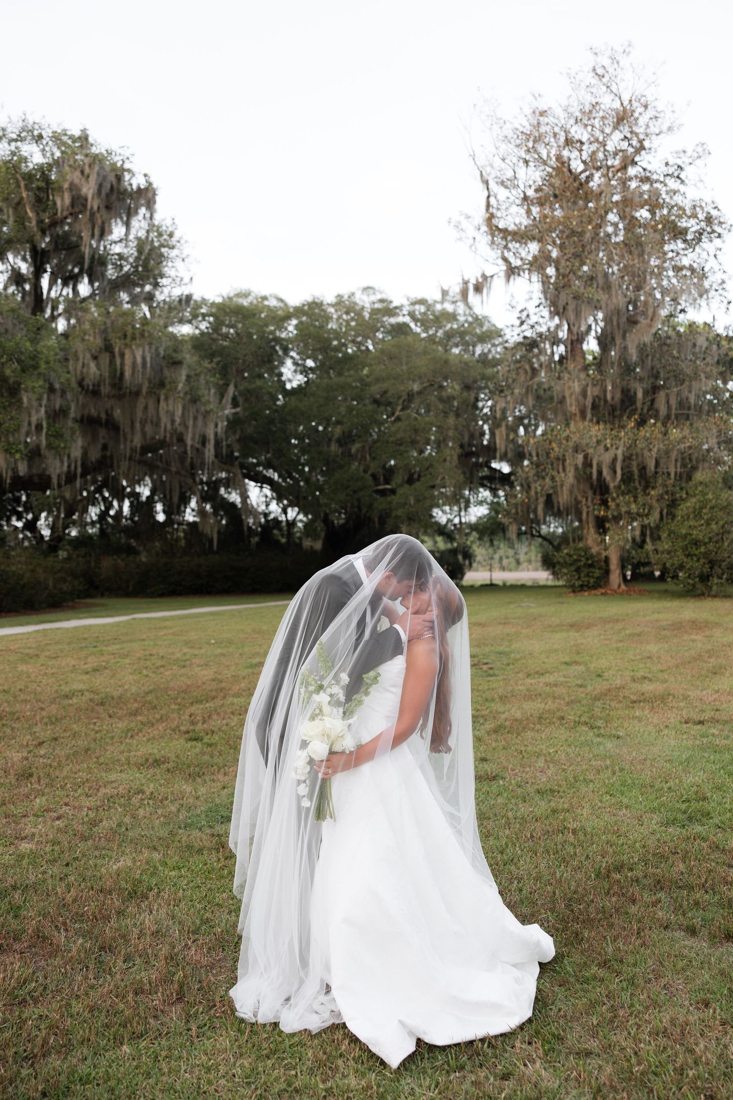 Bride in white gown and flowing veil standing in grassy field with Spanish moss trees in background.