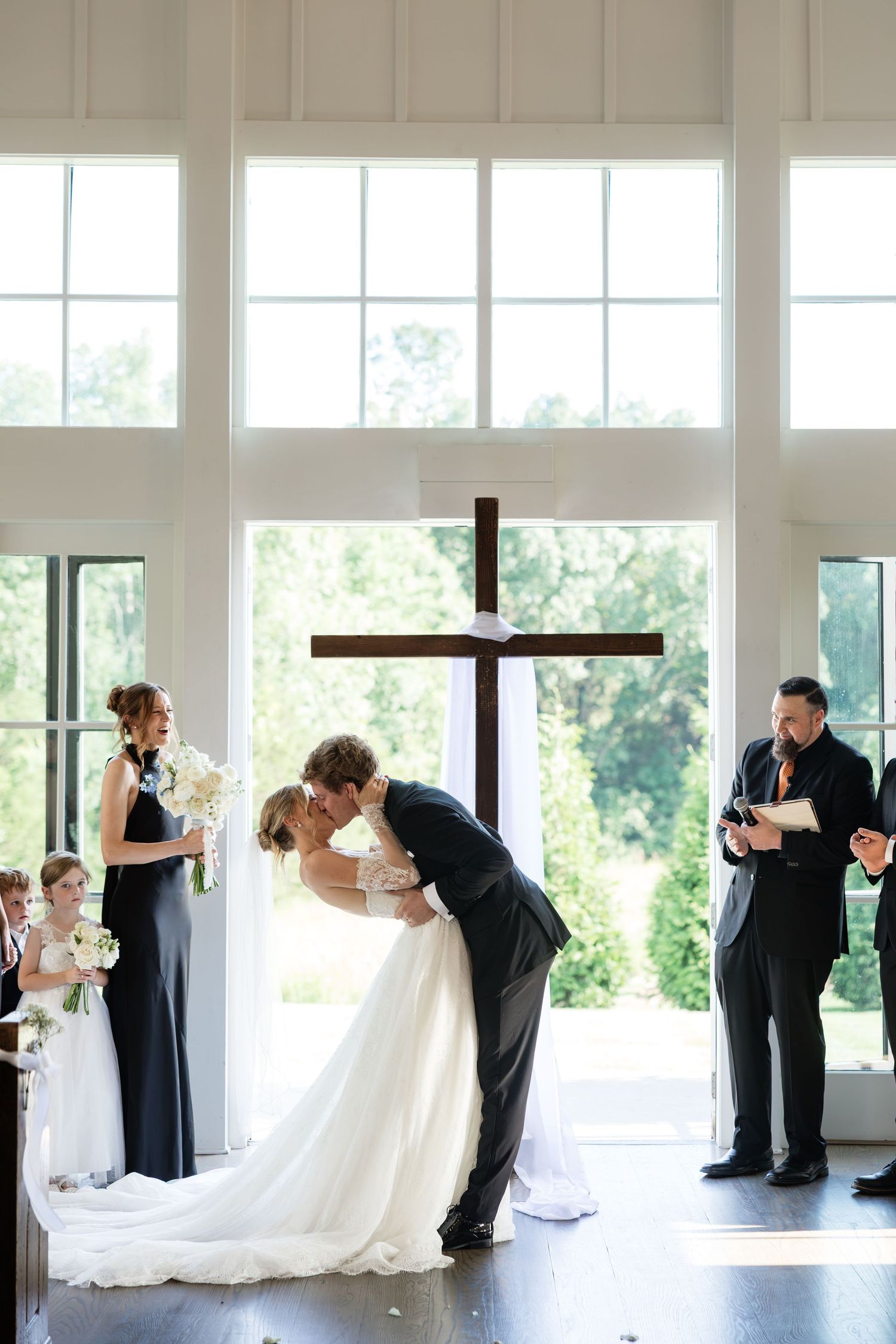 Bride and groom share first kiss at wedding altar with wooden cross in bright chapel with large windows.
