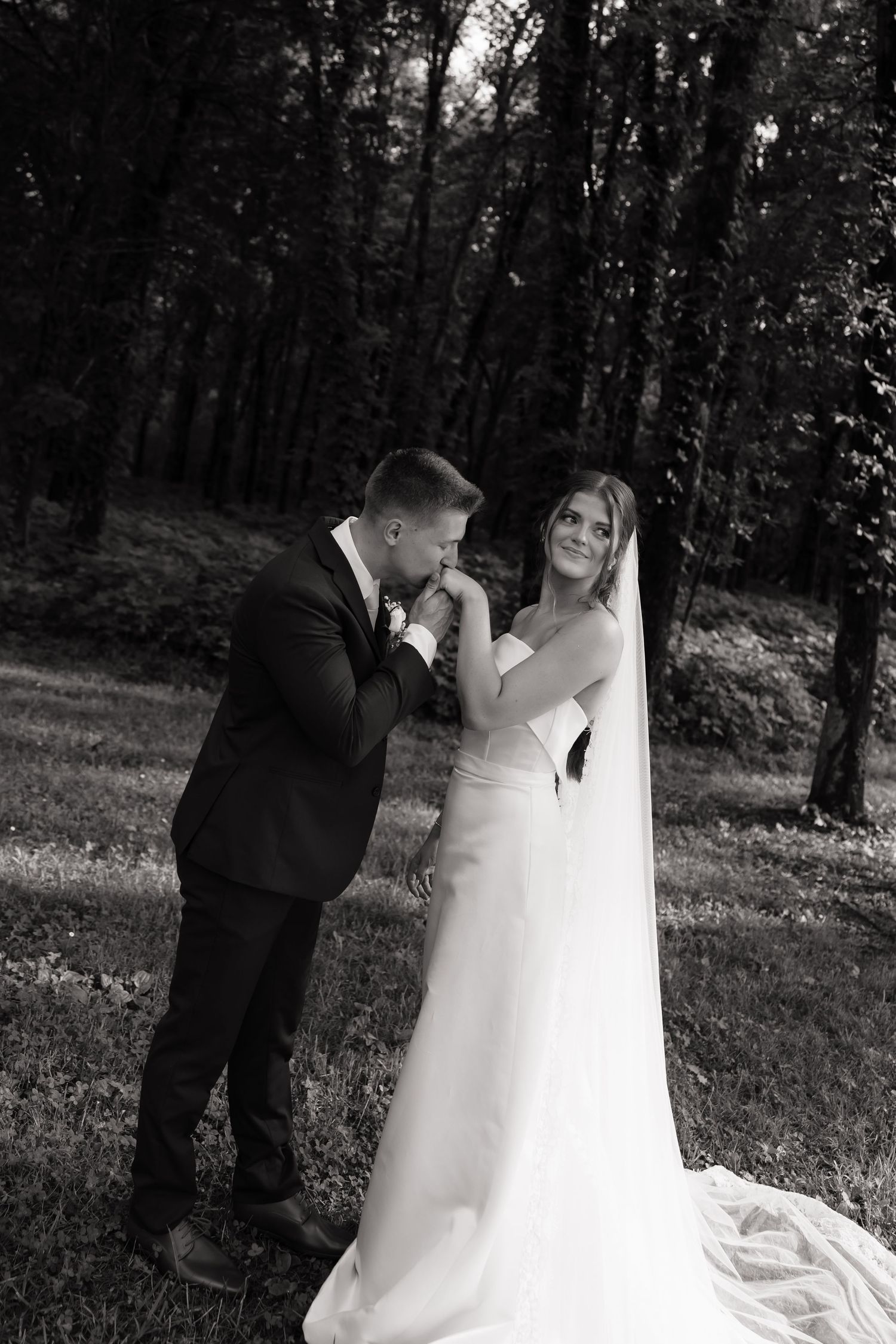 Groom kisses bride's hand in a romantic black and white wedding portrait surrounded by lush forest trees.