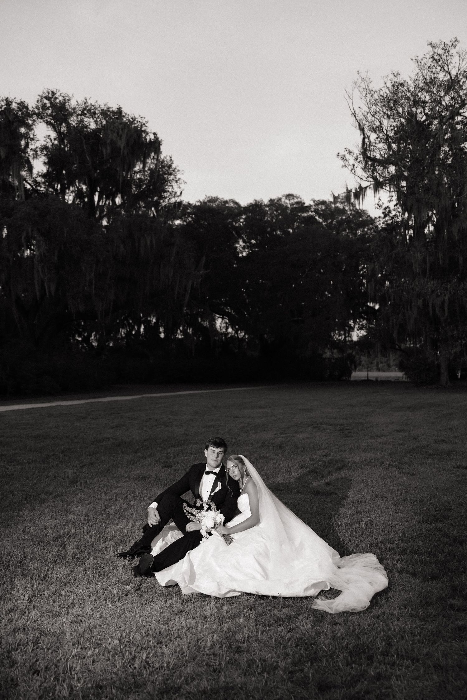 Black and white photo of bride and groom sitting on lawn surrounded by moss-draped trees at dusk.