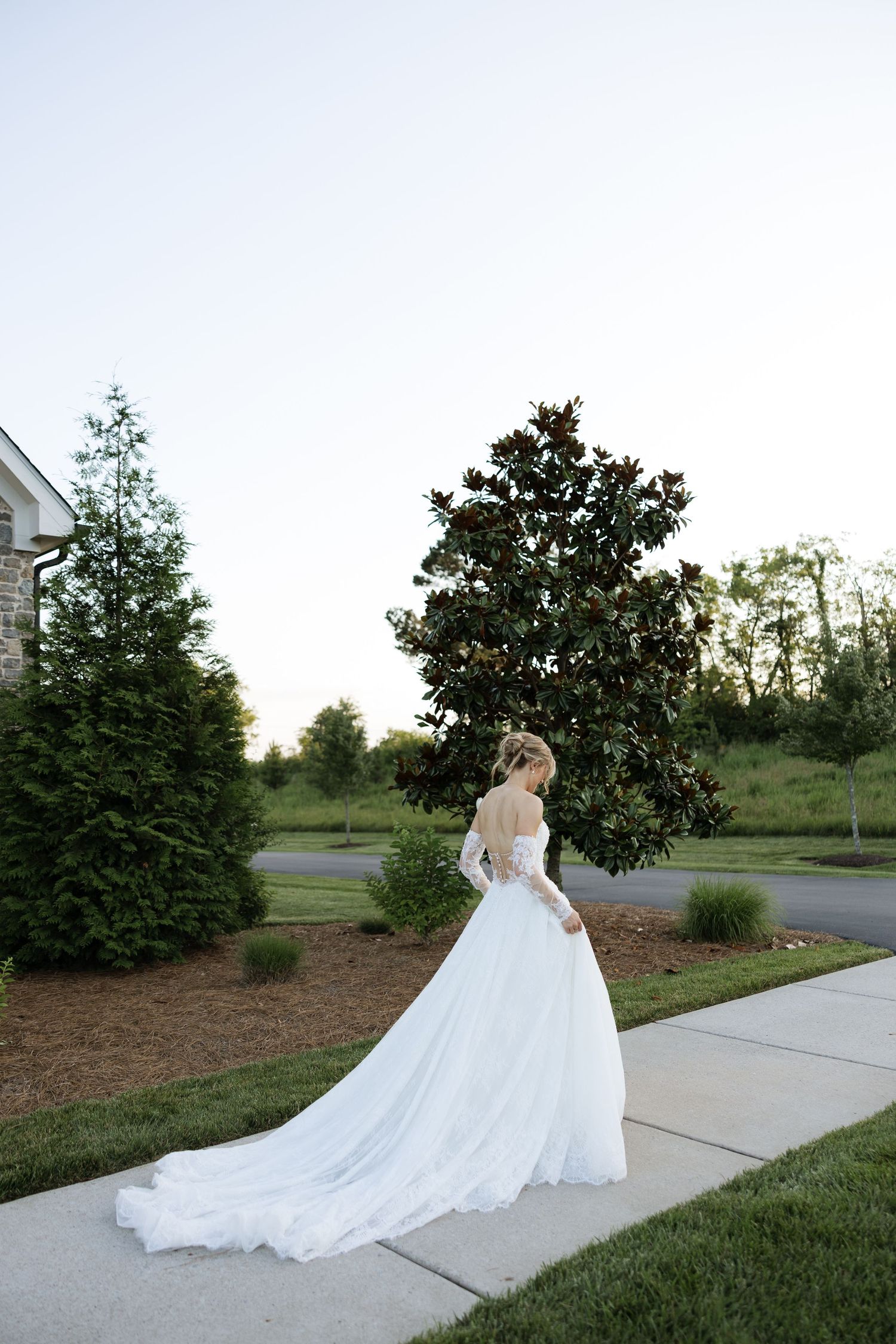 Bride in long-sleeve lace wedding gown with cathedral train stands near magnolia tree at dusk.