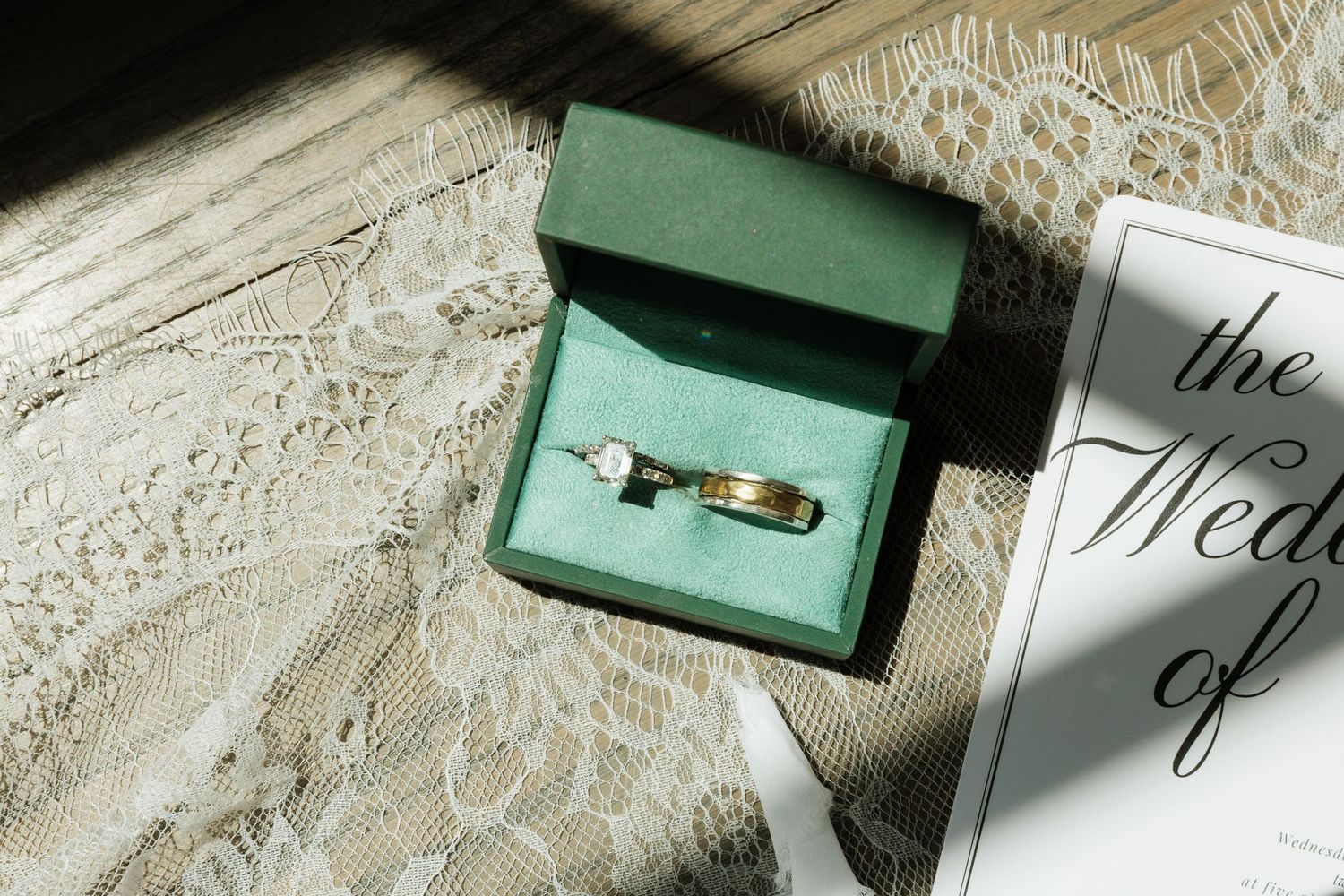 Gold wedding bands and diamond engagement ring displayed in green velvet box beside wedding program on lace fabric.