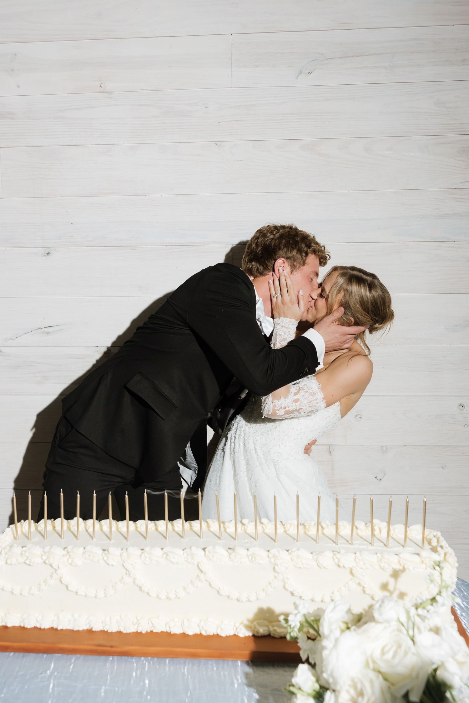 Bride and groom share a kiss over large white sheet wedding cake with gold candles at their reception.