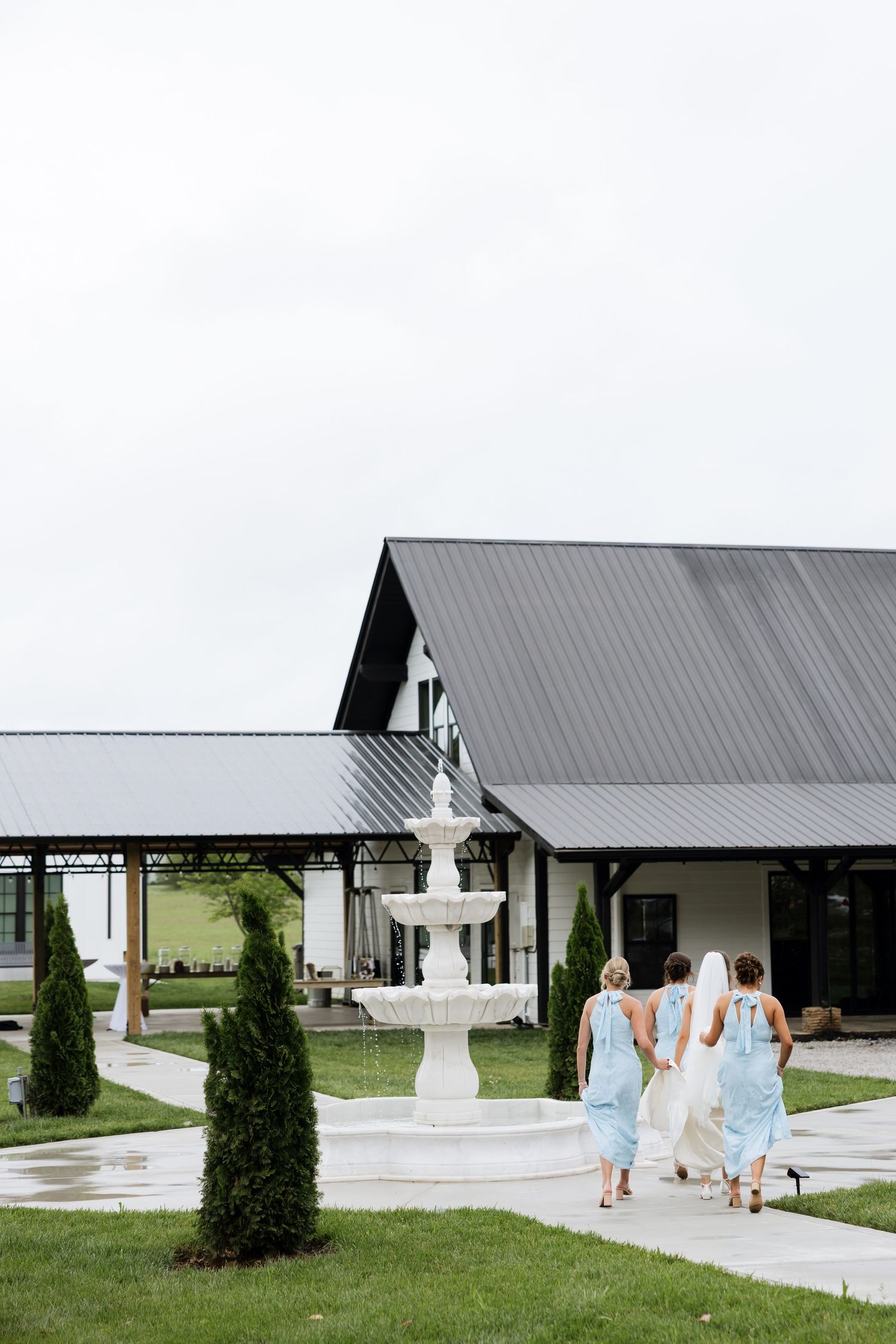 Bride and bridesmaids in light blue dresses walking toward a white barn wedding venue with a tiered fountain.