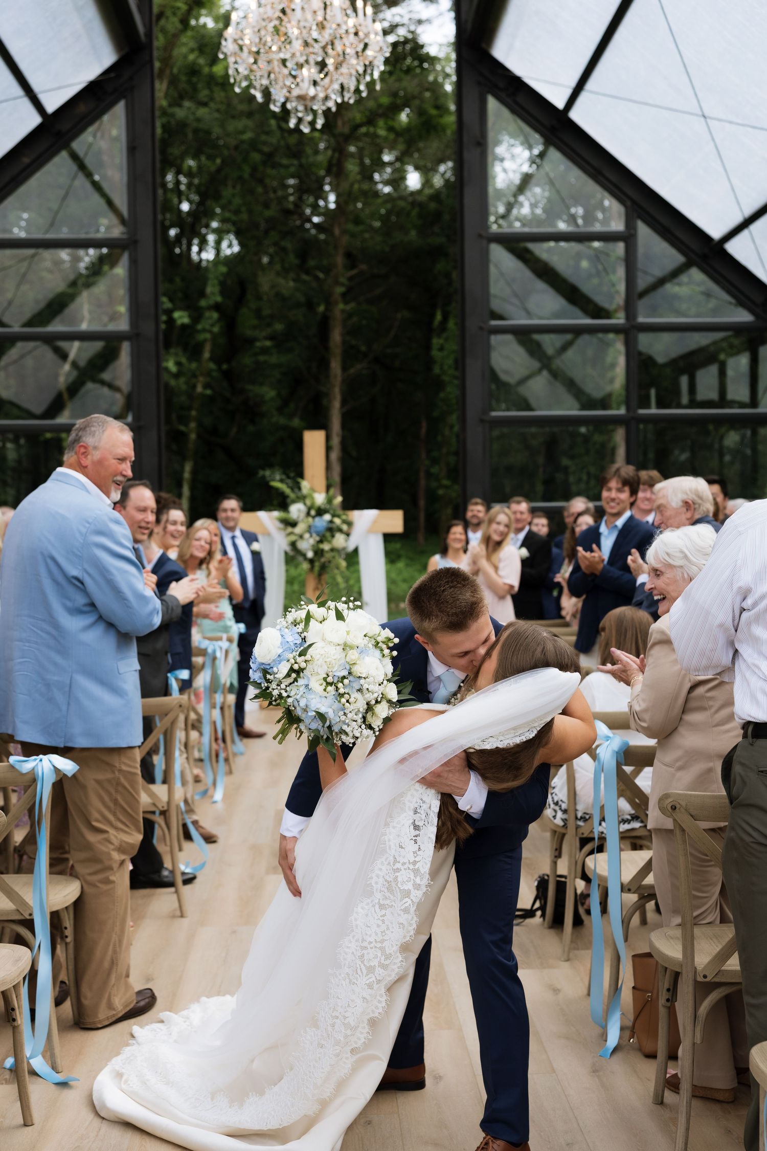 Groom dips bride in aisle of glass chapel as guests applaud during wedding ceremony.