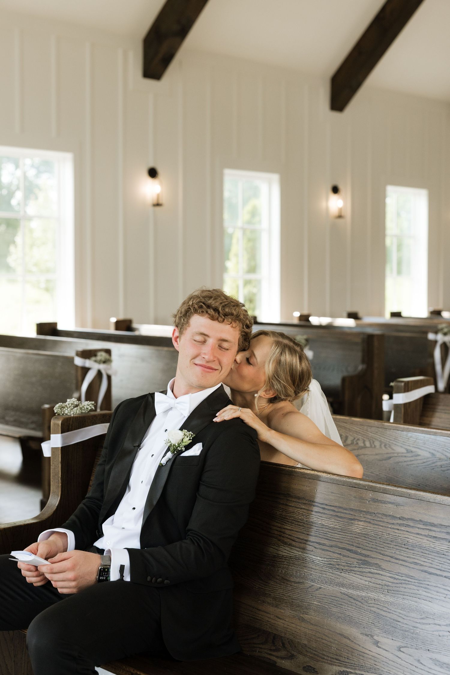 Bride kissing groom on cheek while sitting in wooden church pews during wedding ceremony.