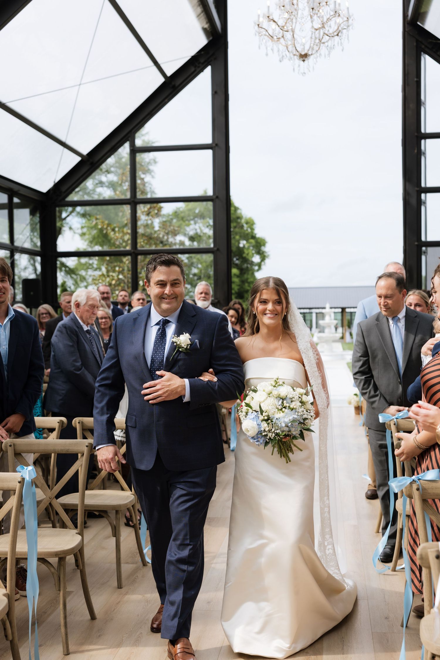 Bride and groom walk down the aisle together in an elegant glass chapel with chandelier and blue ribbon chairs.