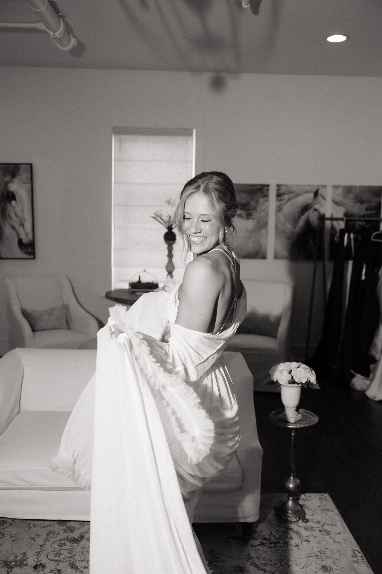 Bride in elegant backless wedding gown smiling while holding her dress train in a bridal suite, black and white photo.