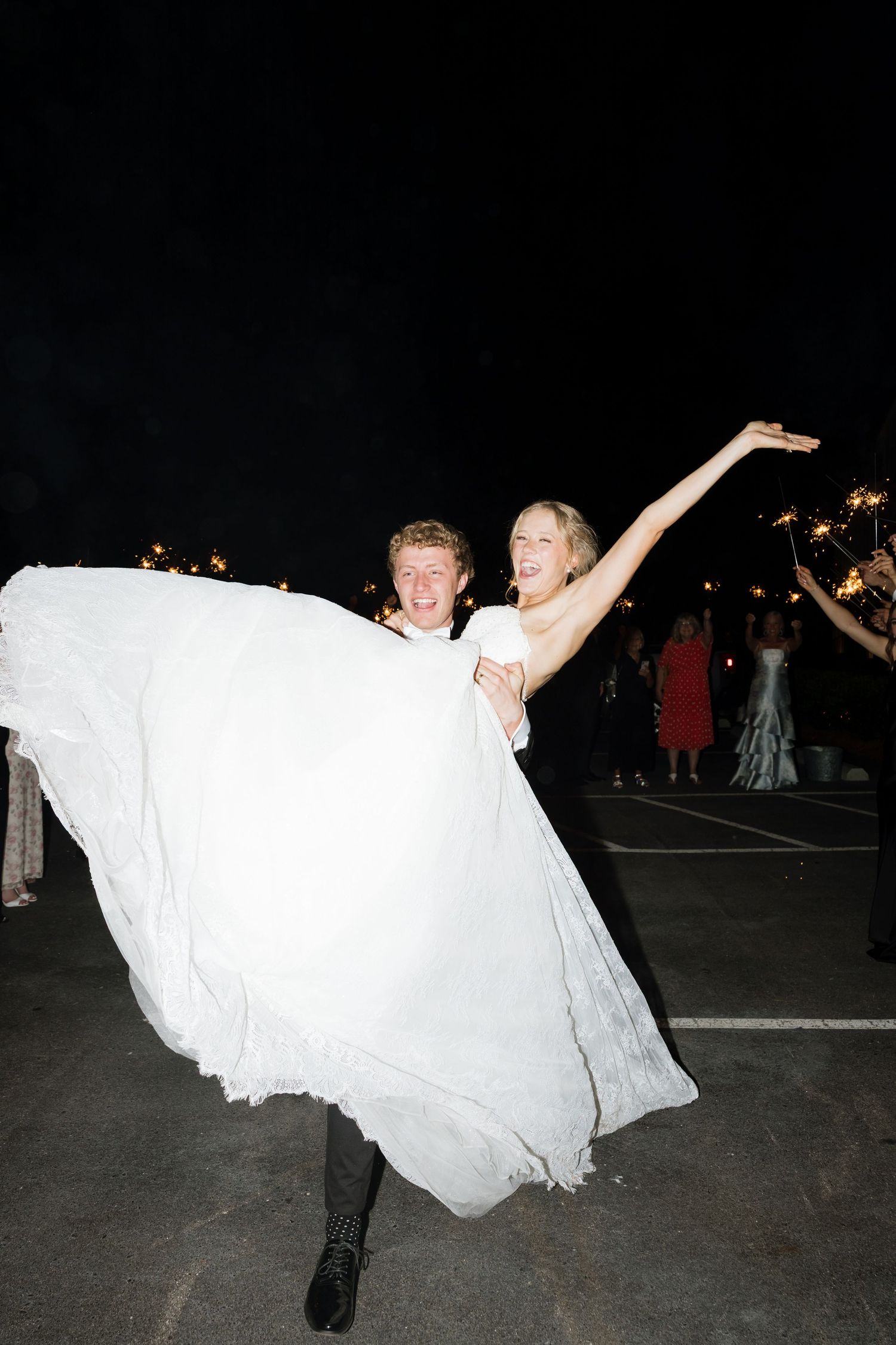 Groom lifting joyful bride in white wedding gown during outdoor nighttime sparkler sendoff celebration.