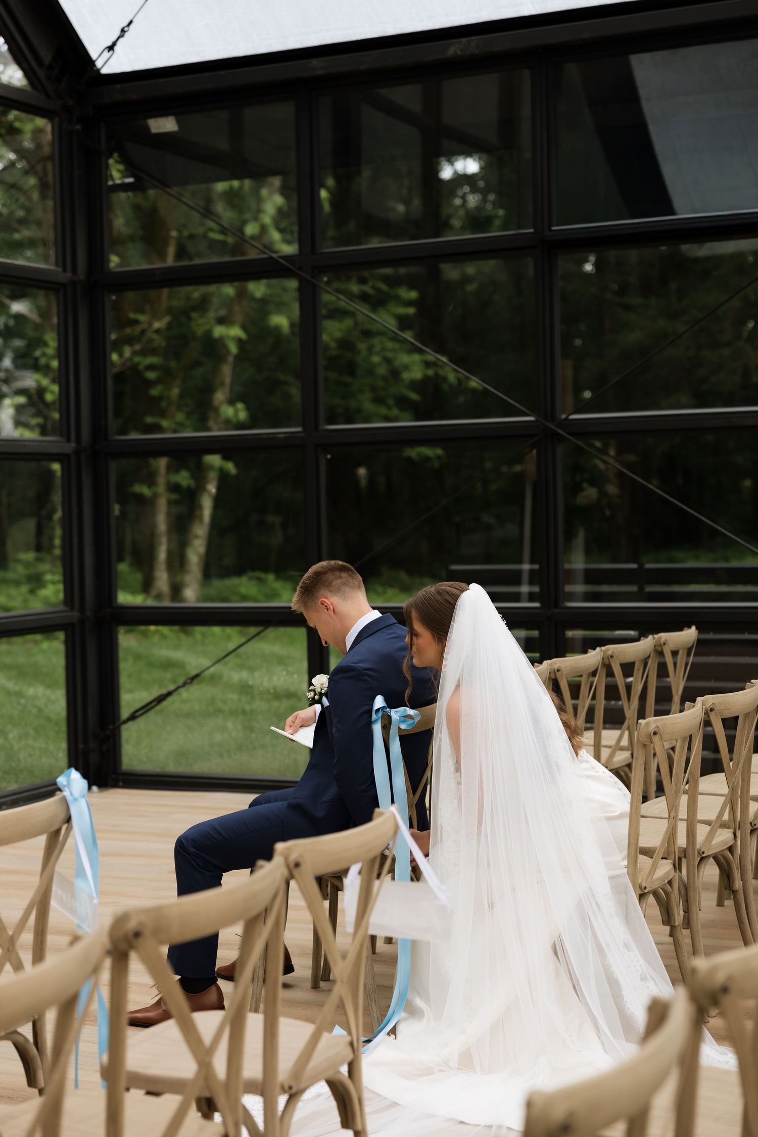 Bride and groom sitting together in wooden chairs inside a glass greenhouse wedding venue surrounded by lush greenery.