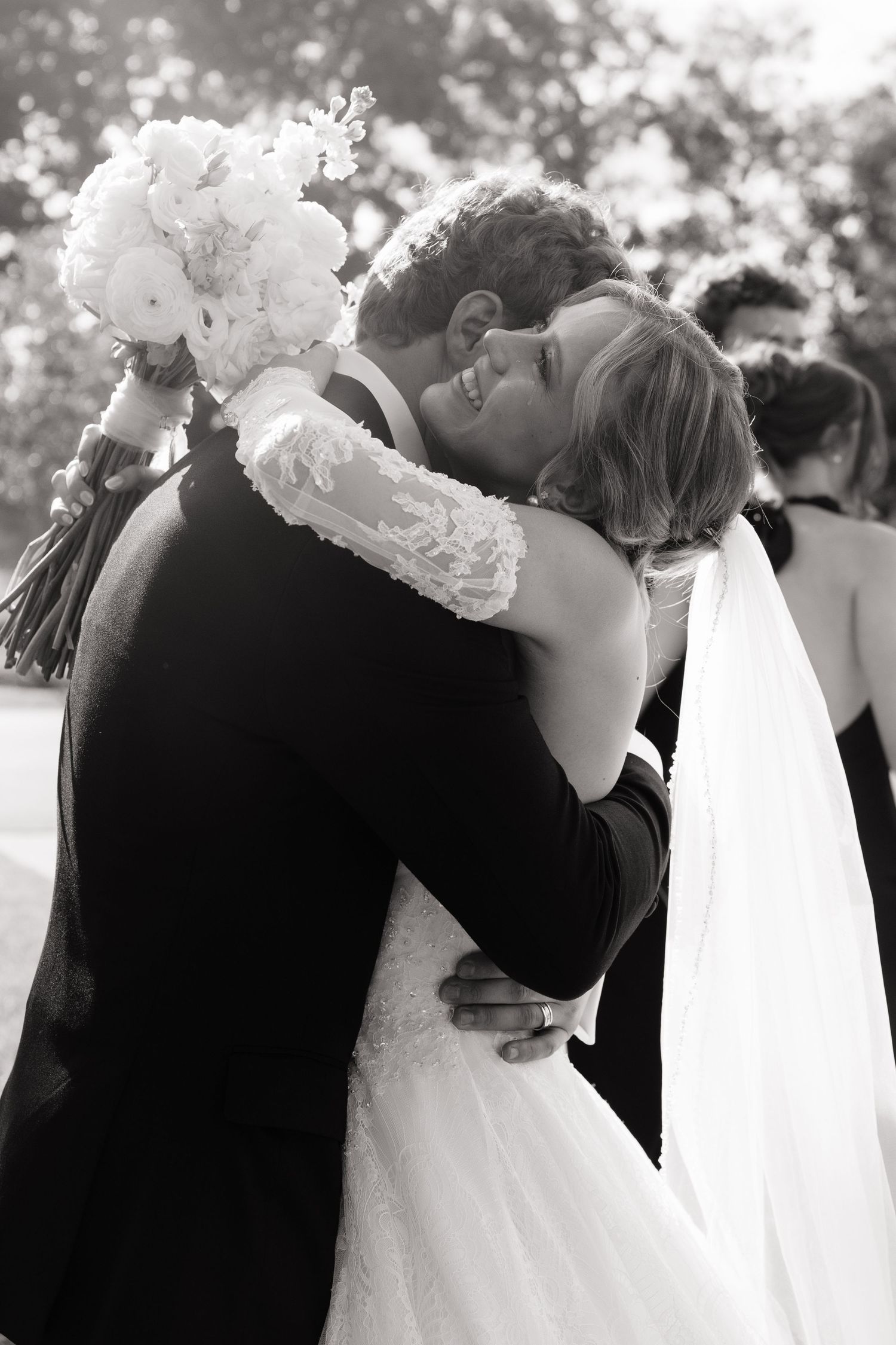 Black and white photo of a bride and groom sharing a joyful embrace, with the bride holding a white floral bouquet.