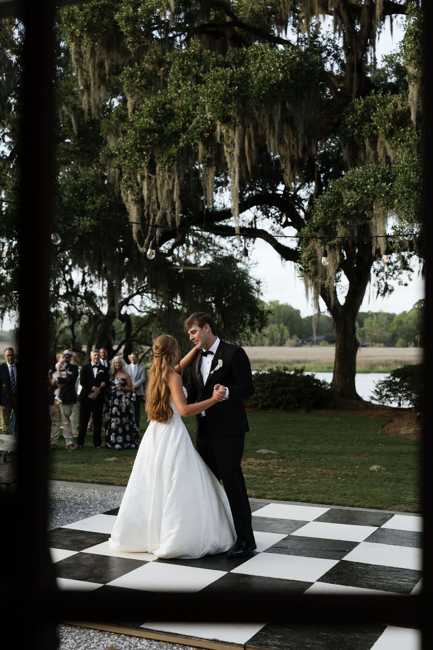 Bride and groom share first dance on black and white checkered floor under Spanish moss-draped oak trees at outdoor wedding.