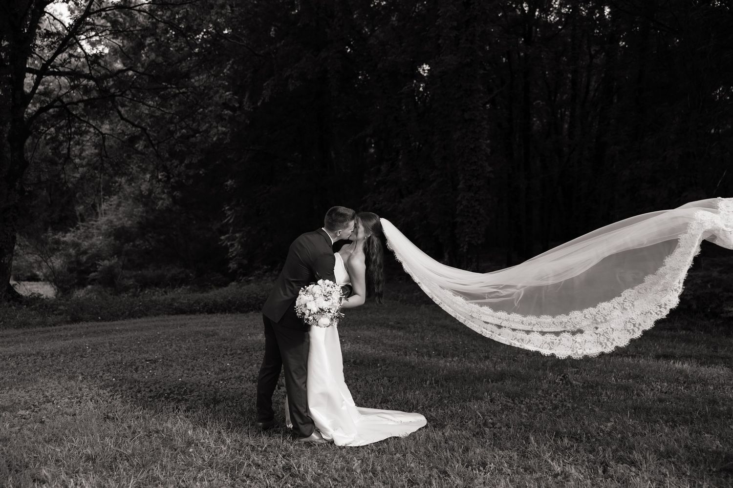 Black and white photo of bride and groom kissing outdoors as her lace veil billows dramatically in the wind.