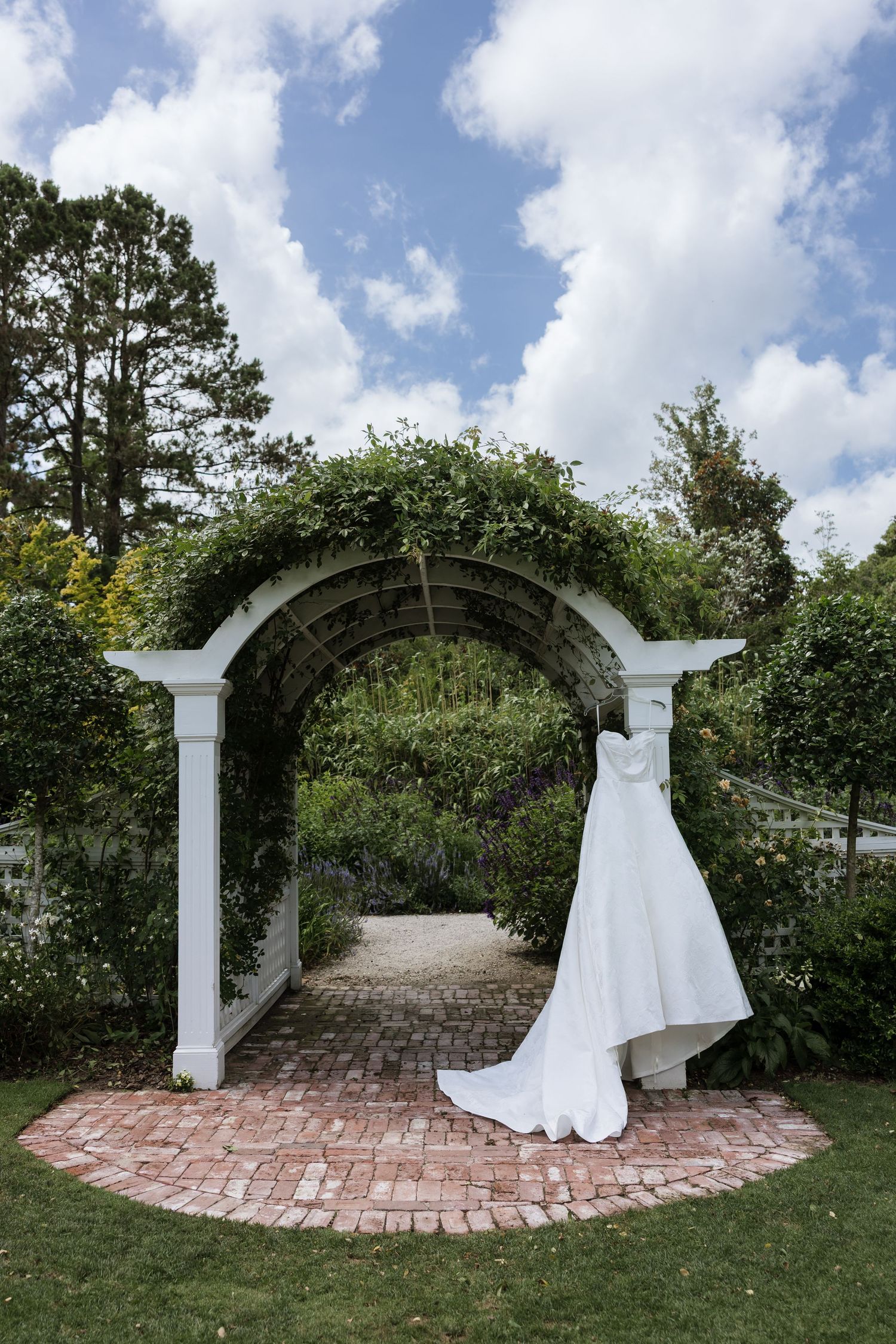 White wedding dress hanging on a vine-covered garden arch with brick pathway and lush greenery under a blue sky.
