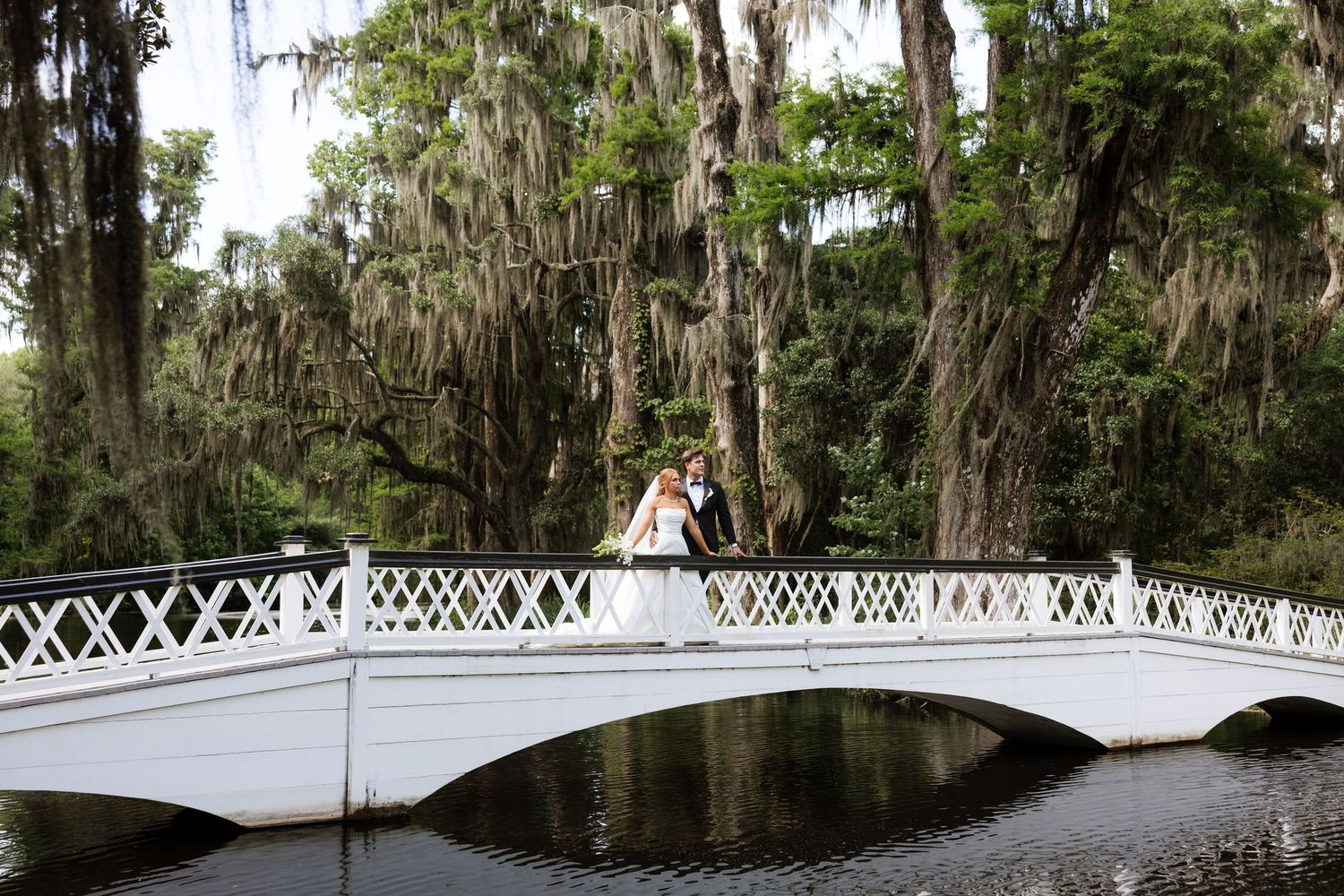 Bride and groom stand on a white ornamental bridge surrounded by moss-draped oak trees reflecting on still water.