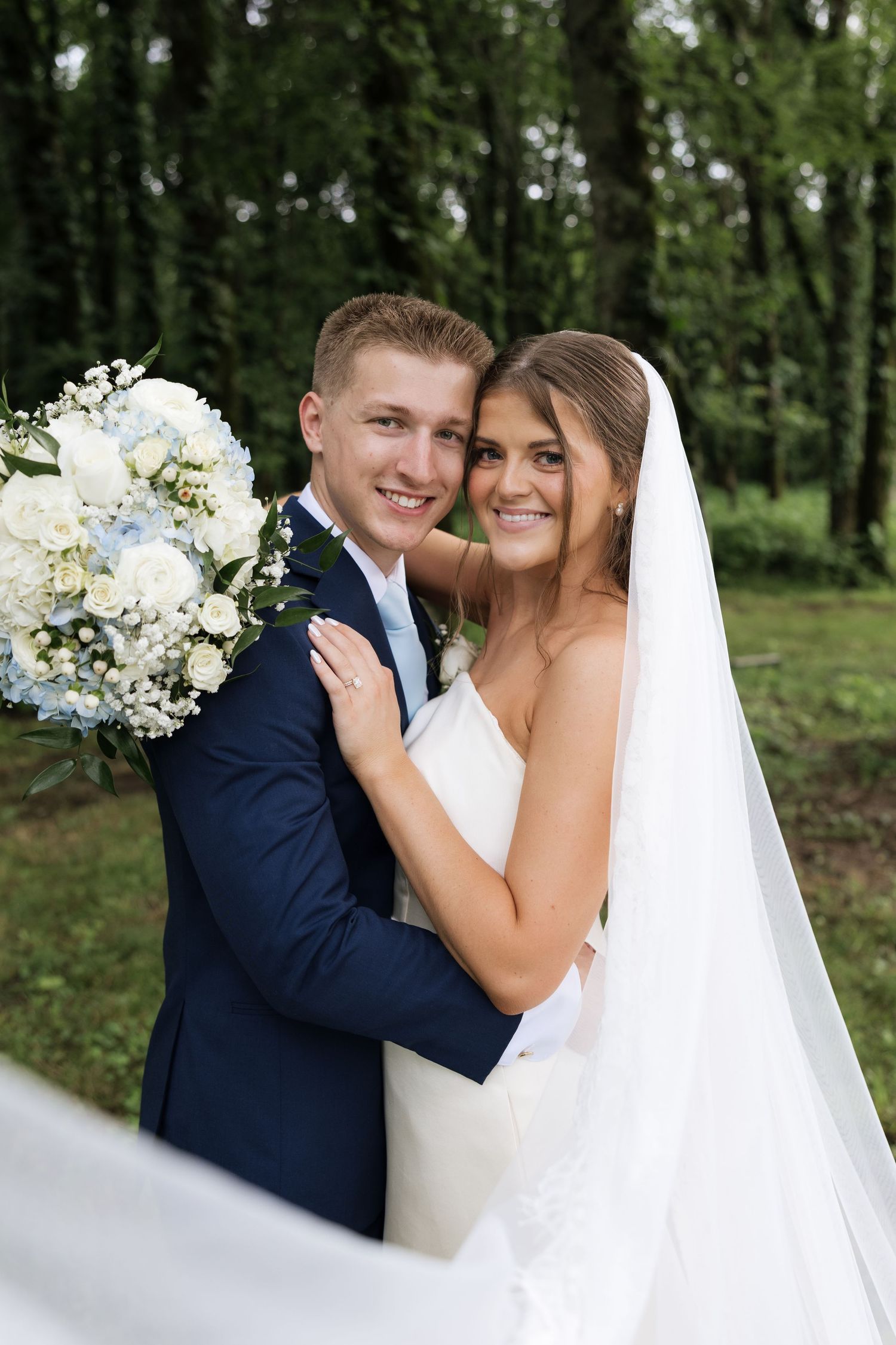 Smiling bride and groom embracing outdoors holding blue and white wedding bouquet.