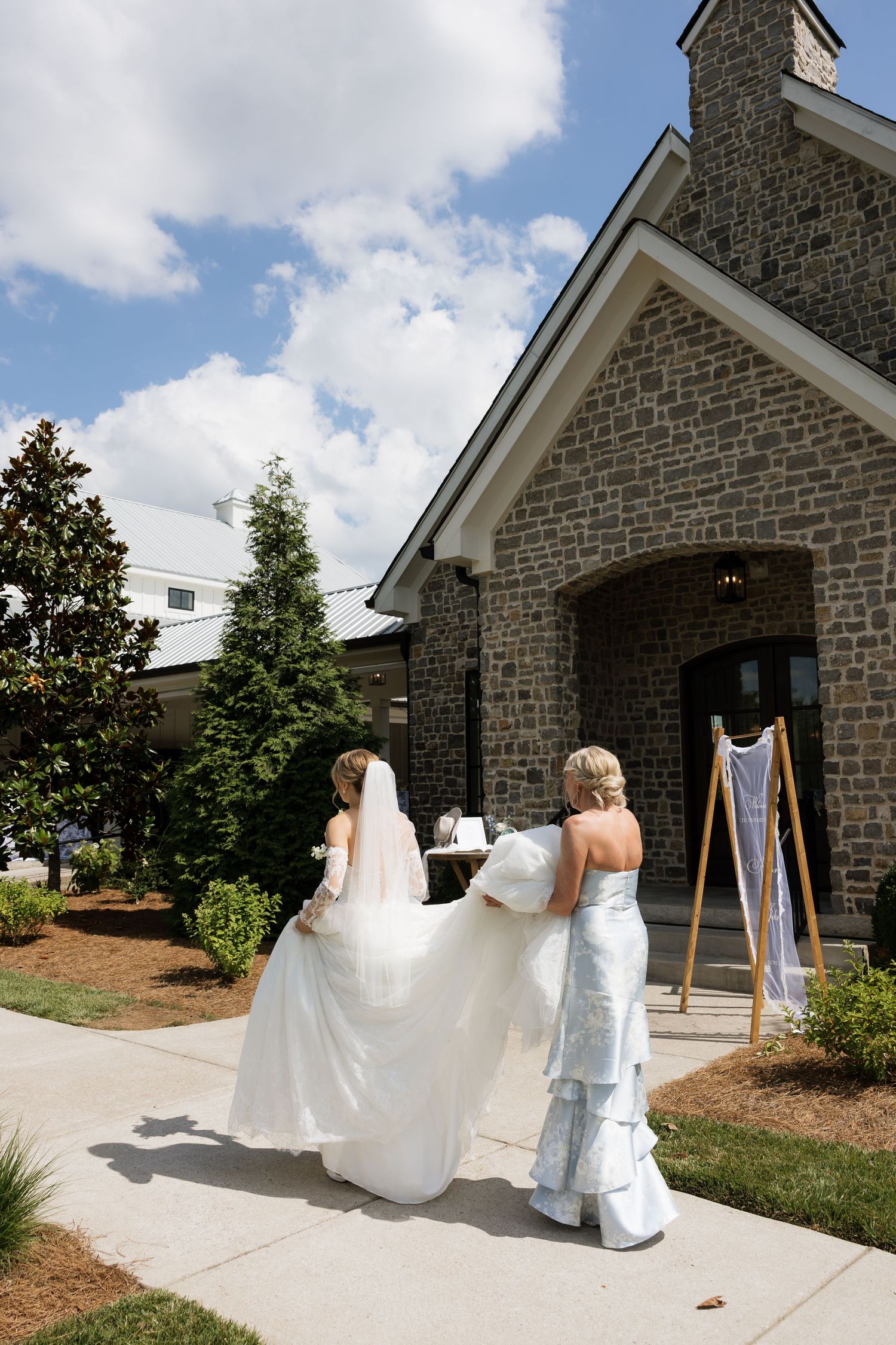 Bride in white gown and veil walks toward stone chapel venue assisted by bridesmaid in blue dress on sunny wedding day.