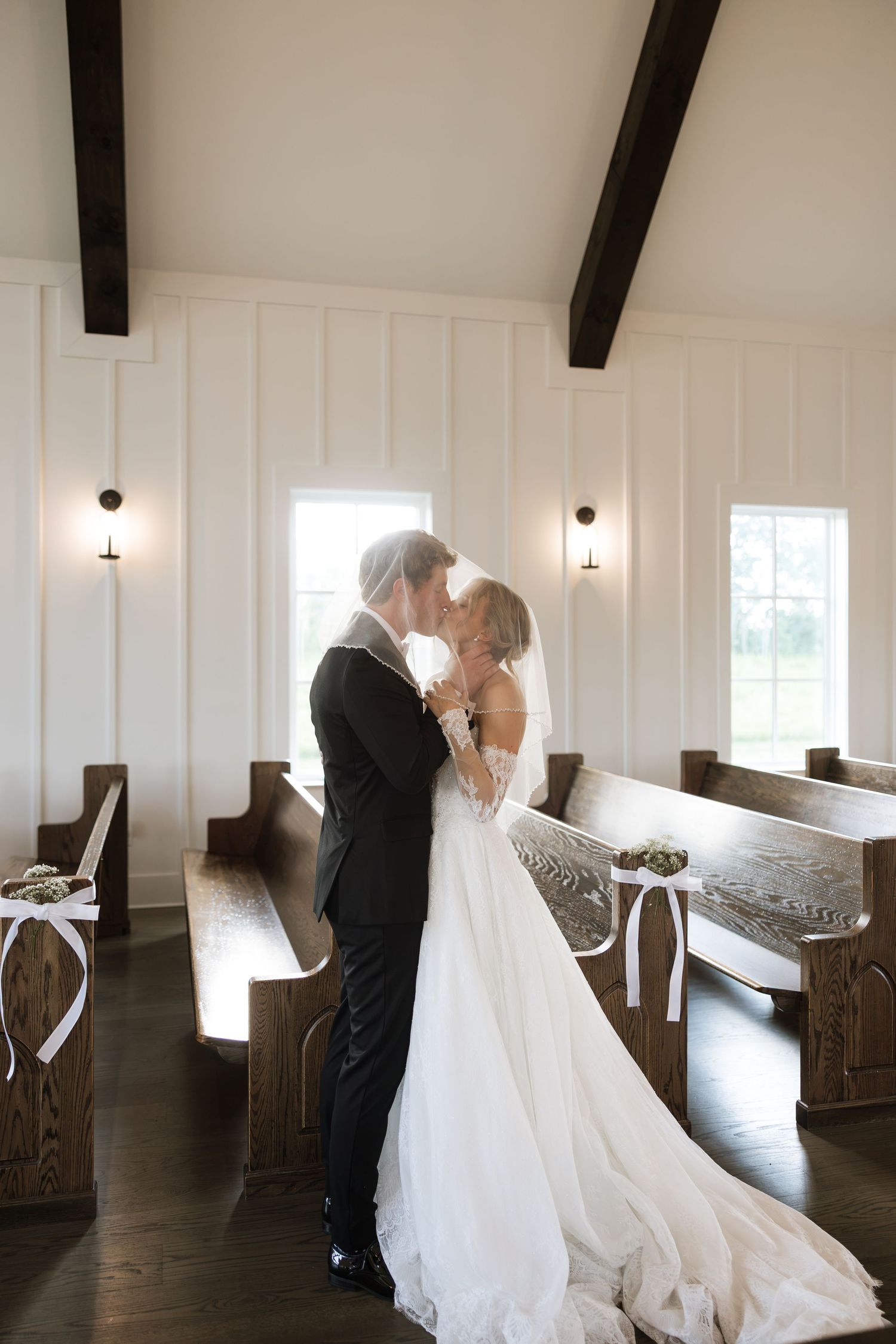 Bride and groom share a kiss in a white chapel with wooden pews, dark beams, and elegant wall sconces.