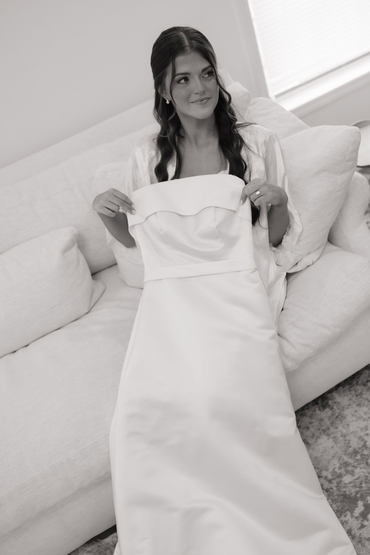 Bride in white strapless wedding gown sitting on couch in black and white photo.