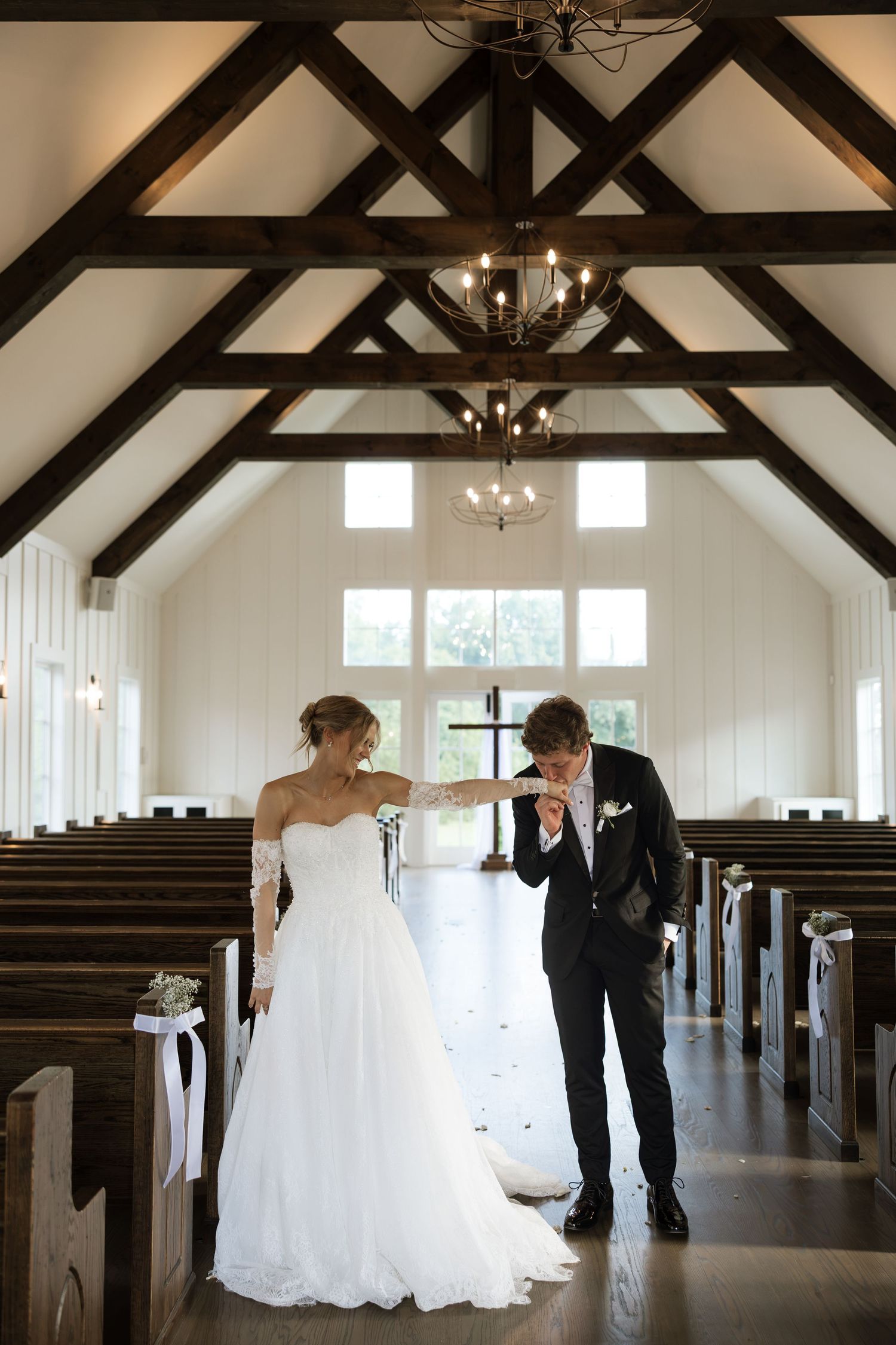 Bride and groom share a tender moment in a white chapel with dark wood beams and chandelier lighting.