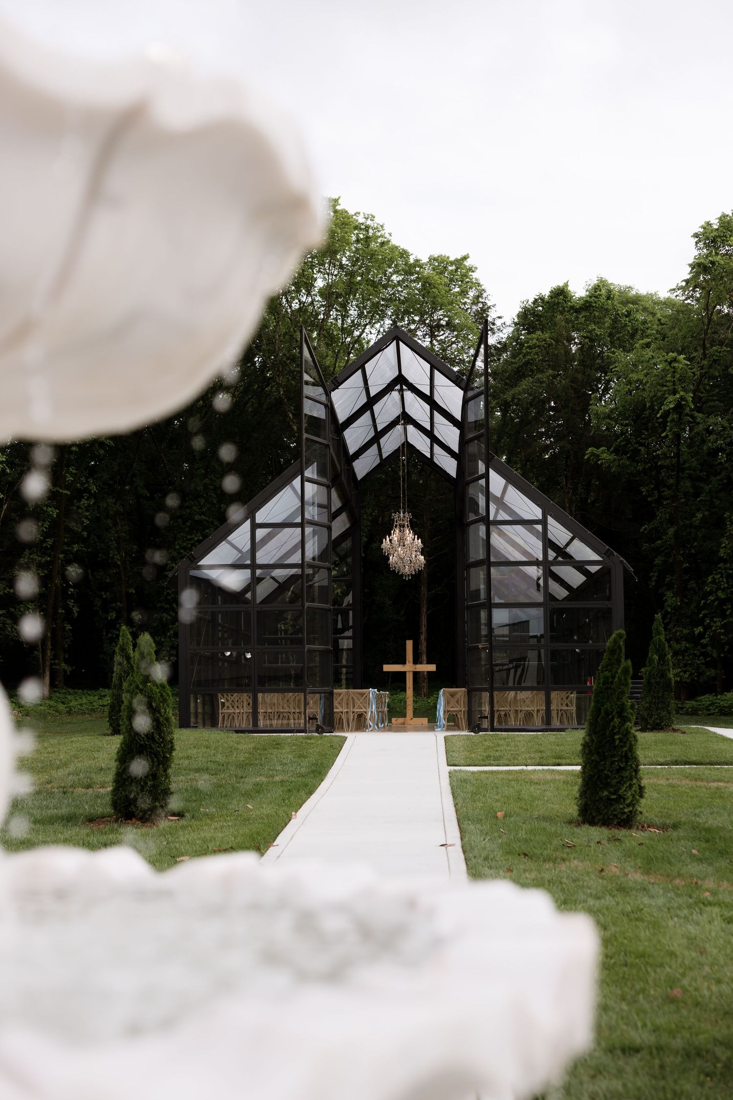 Modern glass chapel with black geometric roof, chandelier, wooden cross, and tree-lined aisle for an elegant outdoor wedding.