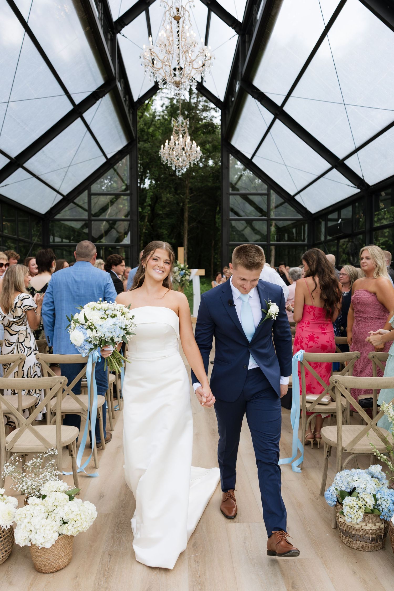 Bride and groom walk down the aisle in a glass greenhouse wedding venue with chandeliers and floral arrangements.