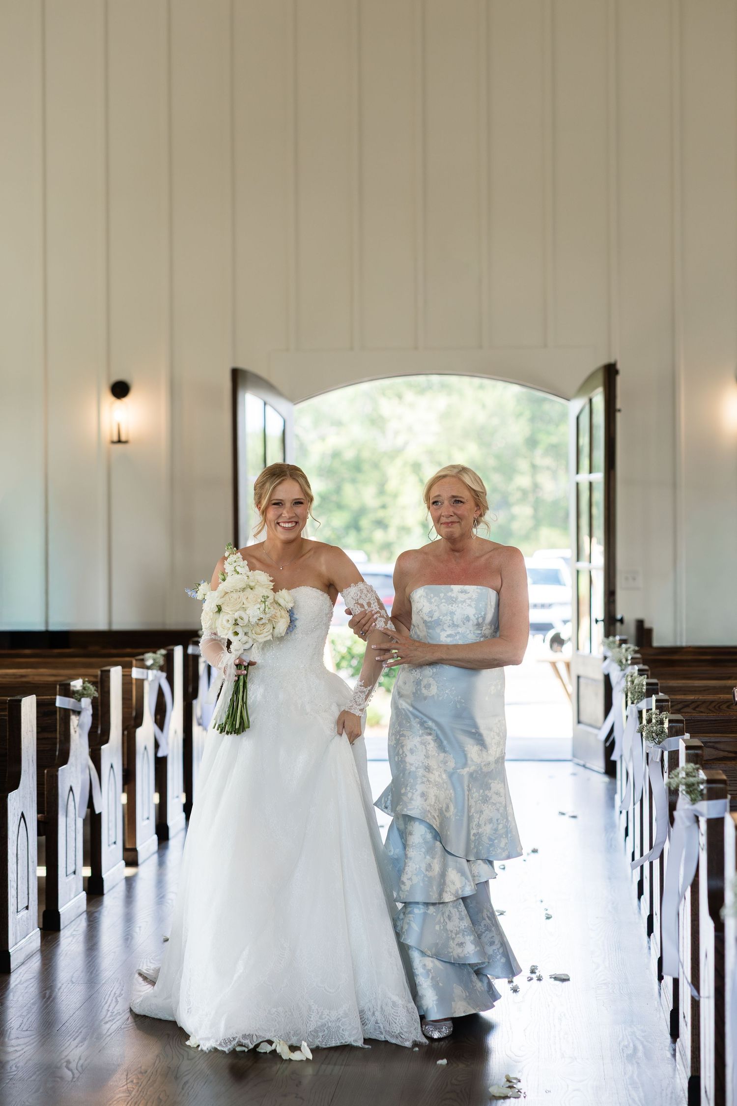 Bride in white strapless gown walks down church aisle with woman in blue floral dress, holding white bouquet.