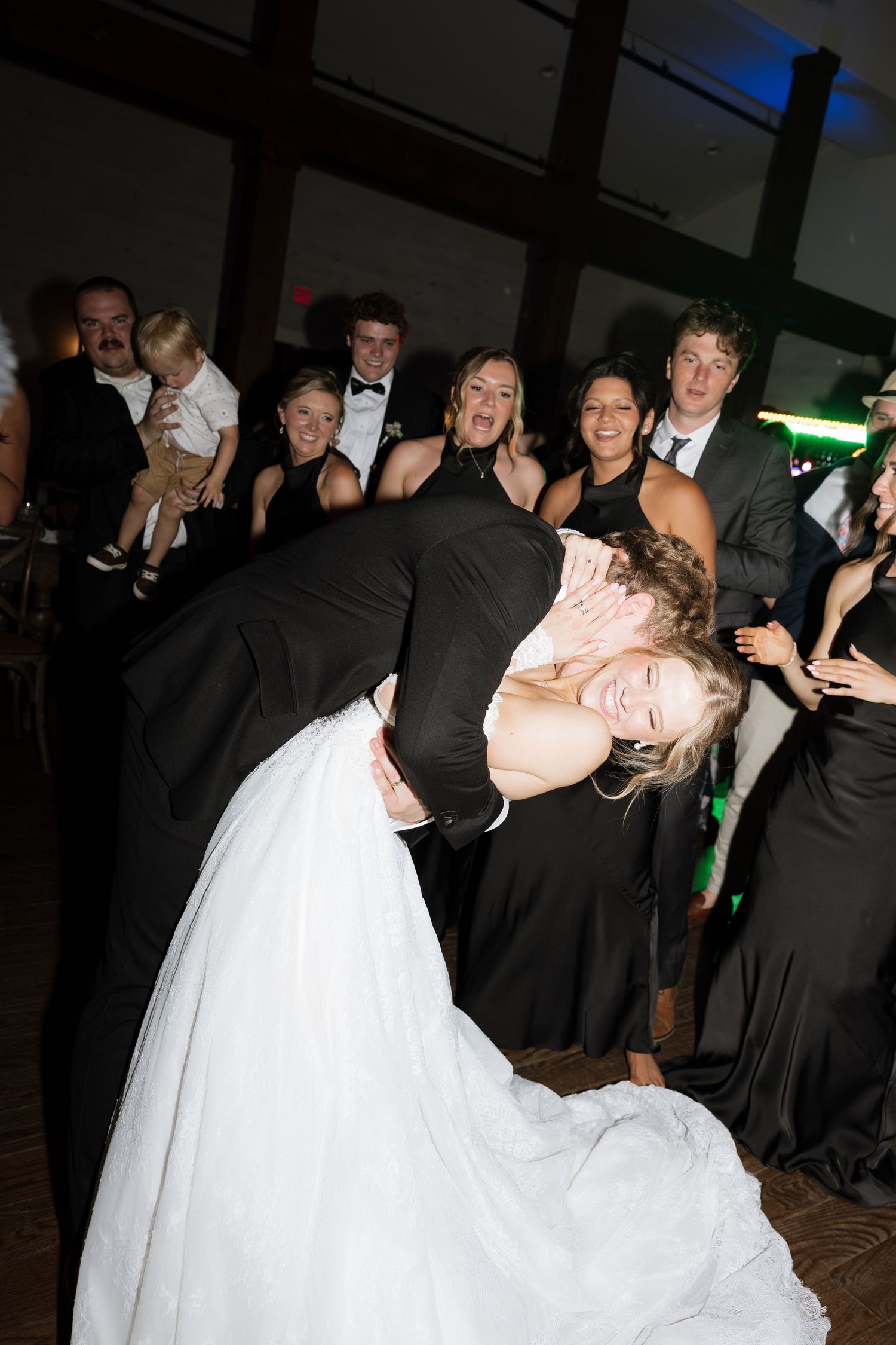 Groom dips laughing bride dramatically on dance floor surrounded by cheering wedding guests during lively reception celebration.
