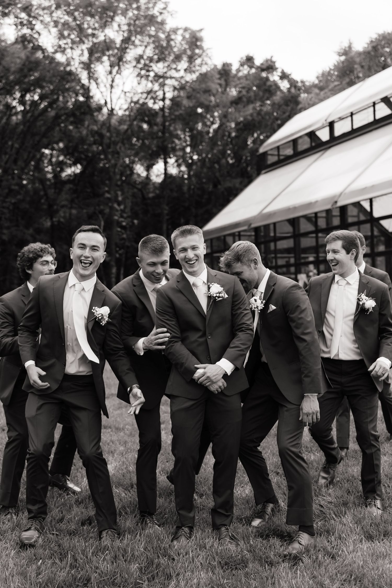 Black and white photo of a groom and groomsmen in dark suits laughing together outdoors near a wedding venue.