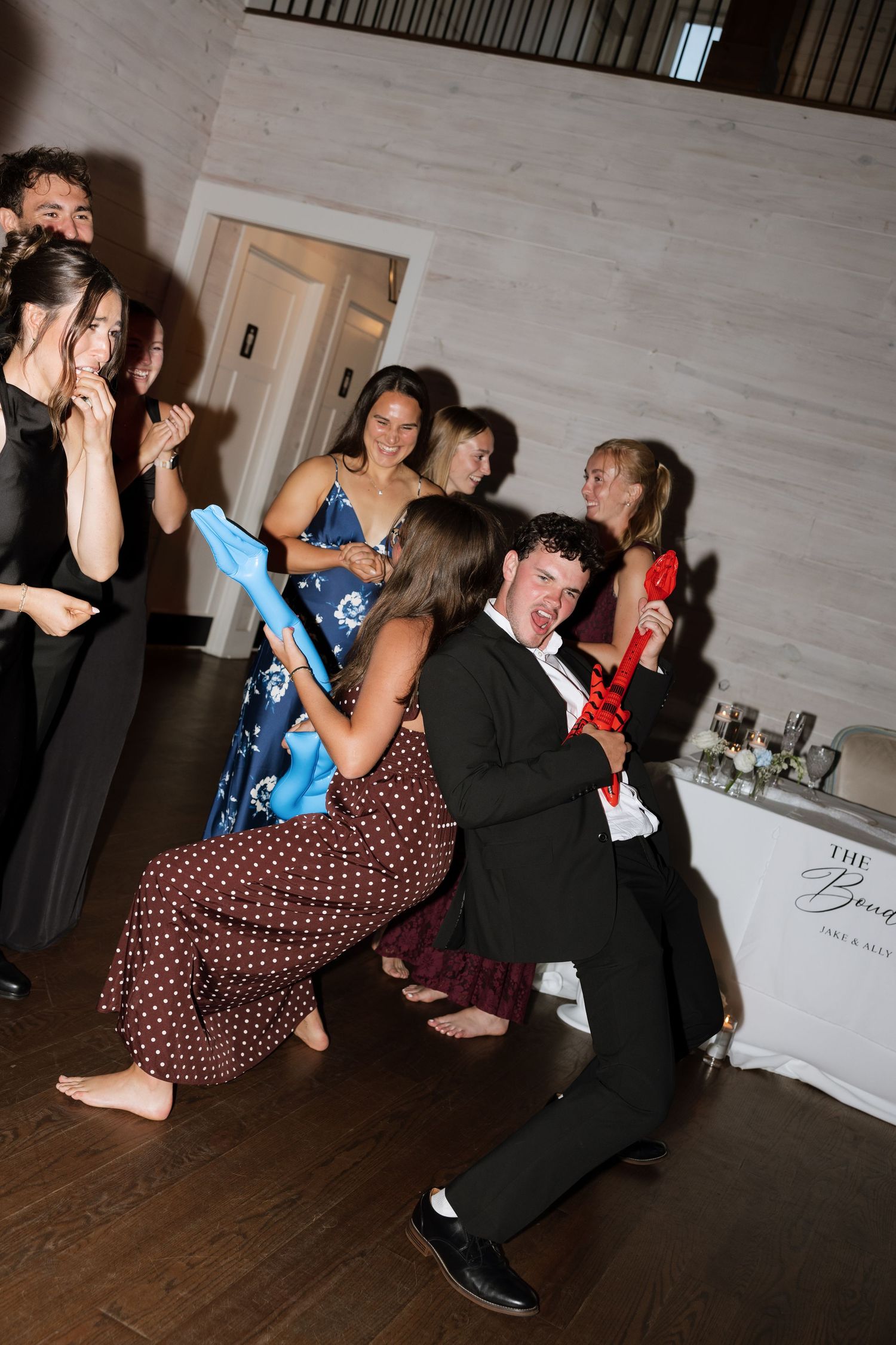 Wedding guests laugh and dance energetically with inflatable guitar at lively reception party.