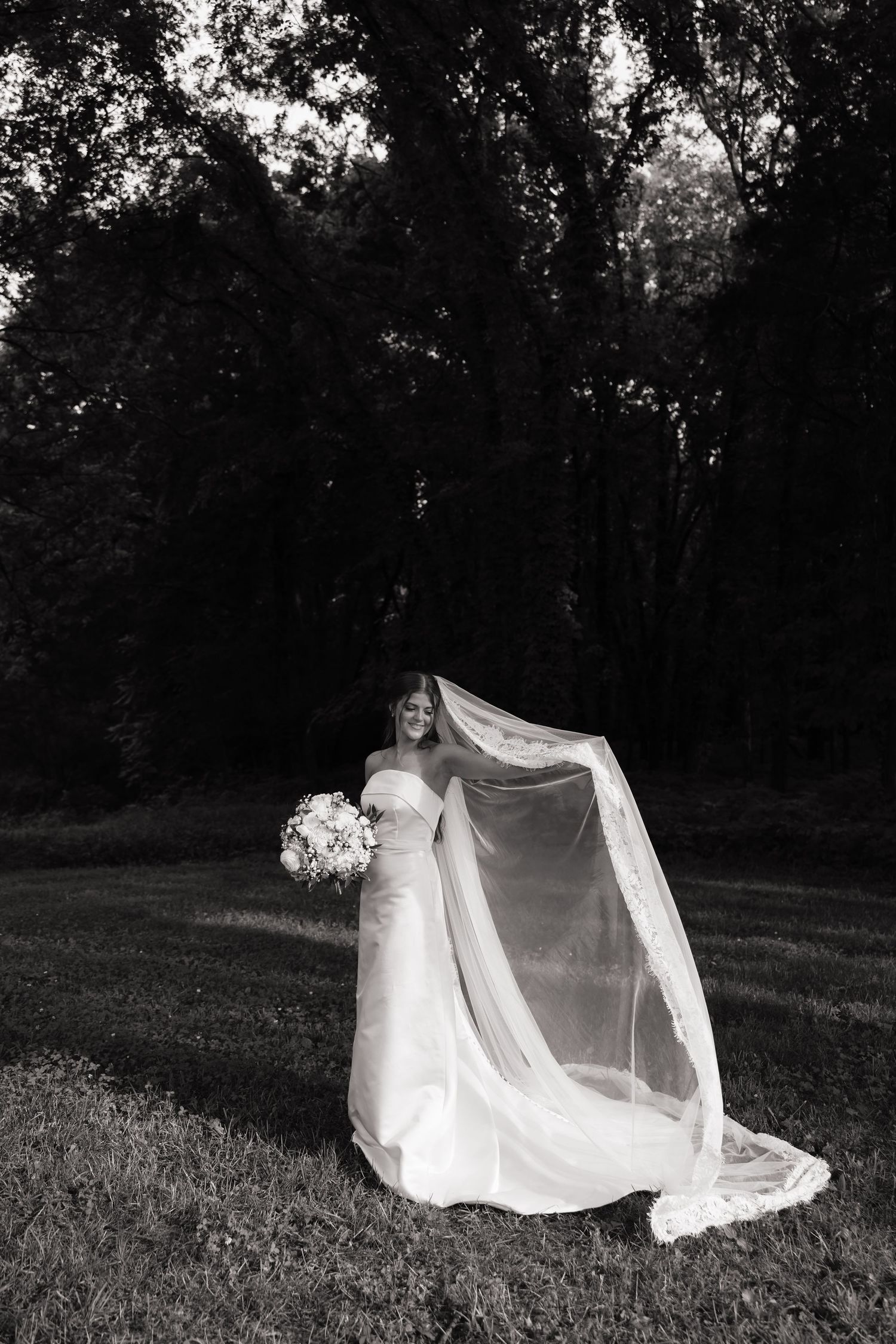 Bride in strapless gown with flowing veil caught in breeze, holding bouquet in black and white outdoor portrait.