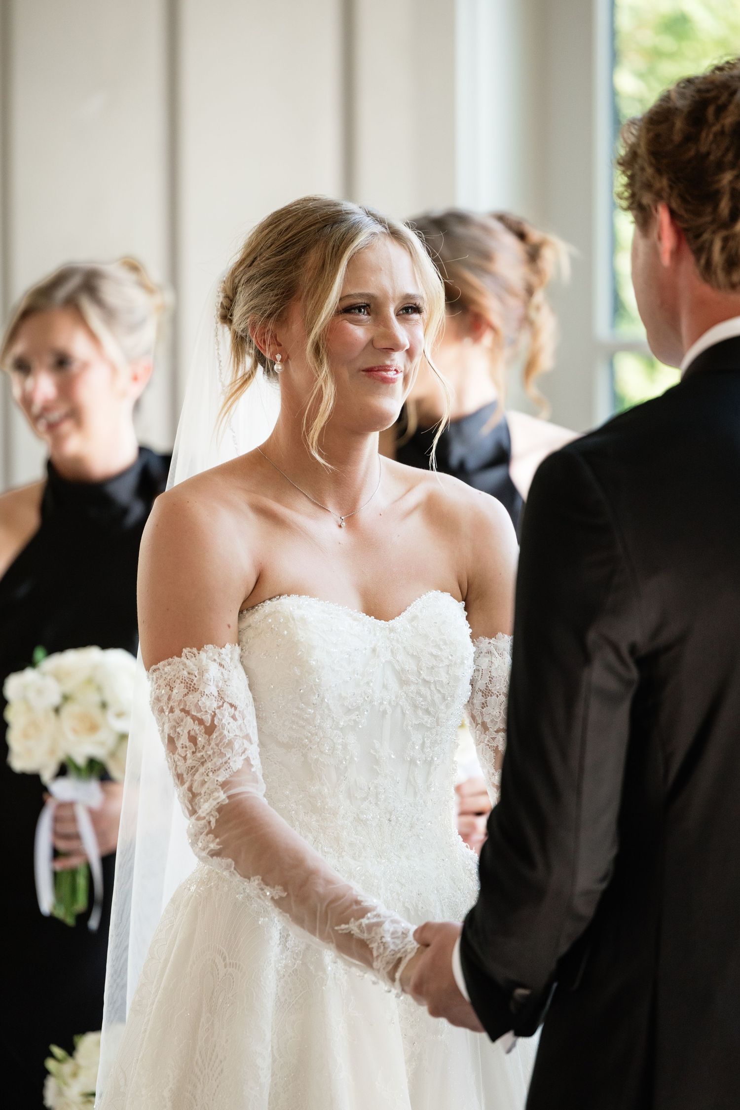 Bride in lace off-shoulder gown holds grooms hands at wedding ceremony, smiling with bridesmaids in black behind.