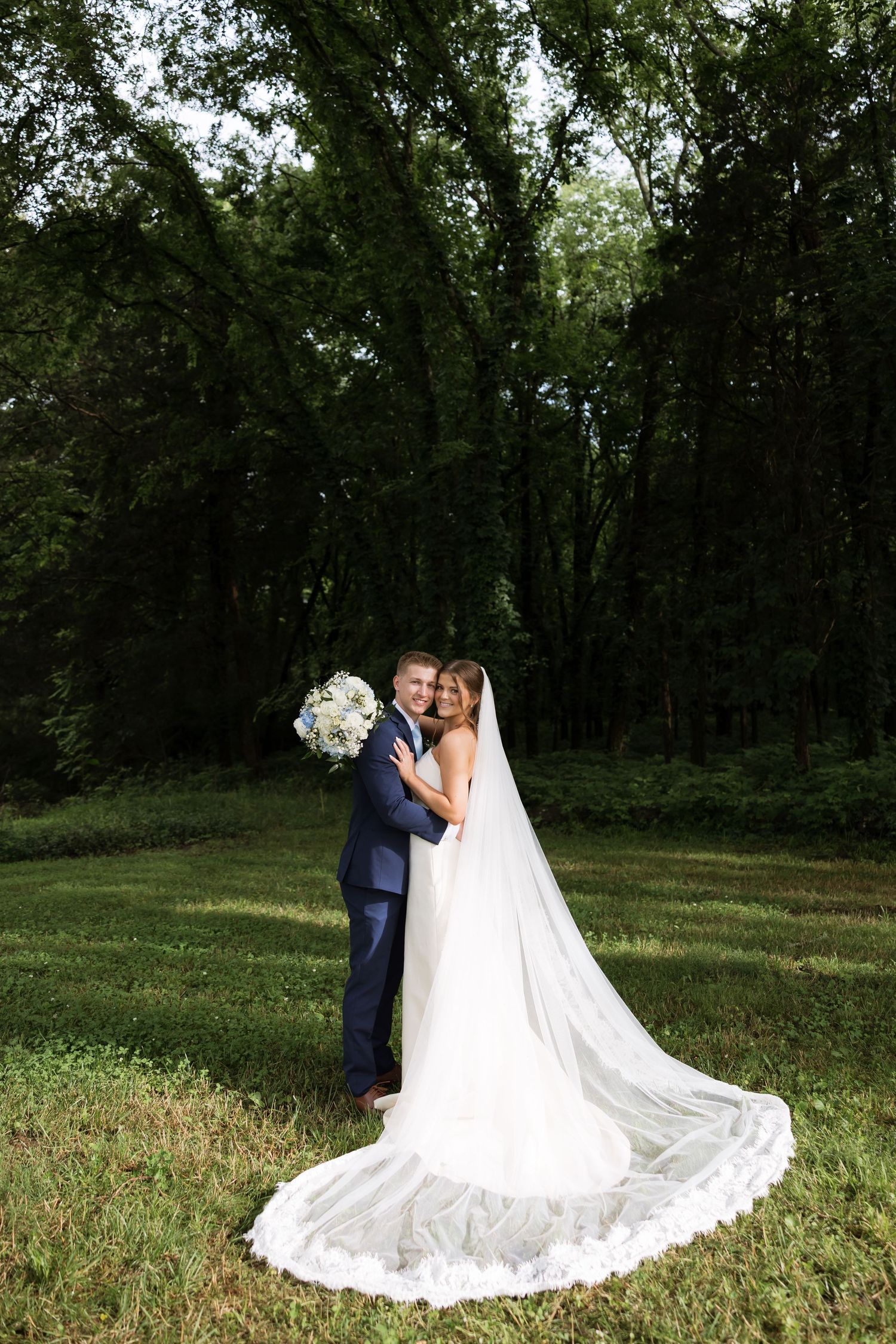 Bride and groom embrace in lush green forest, bride wearing long white gown with cathedral veil and holding white bouquet.