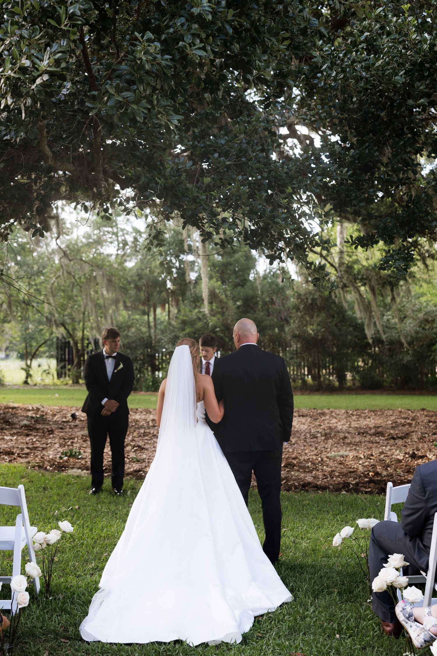 Bride in a flowing white gown and veil walks down the aisle with her father during an outdoor wedding ceremony under a large tree.